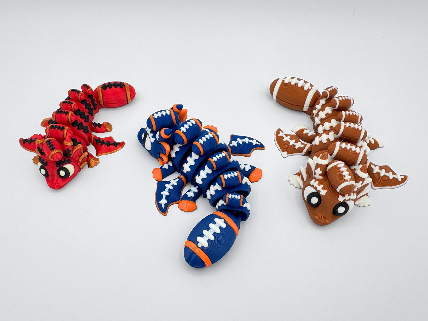 Close-up view of three articulated football dragons in red and gold, blue and orange, and brown and white. Each dragon has detailed football stitching, segmented body for flexible posing, and a football-shaped tail.