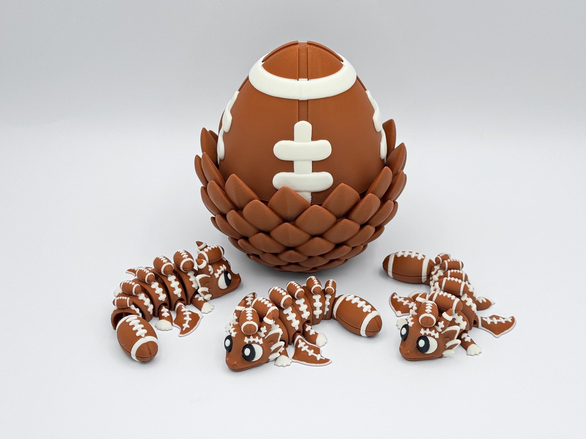 Large brown and white football dragon egg with textured scale base, shown closed. Three small matching articulated dragons with football lace detailing and football-shaped tails are posed in front, all 3D printed in coordinating colors.