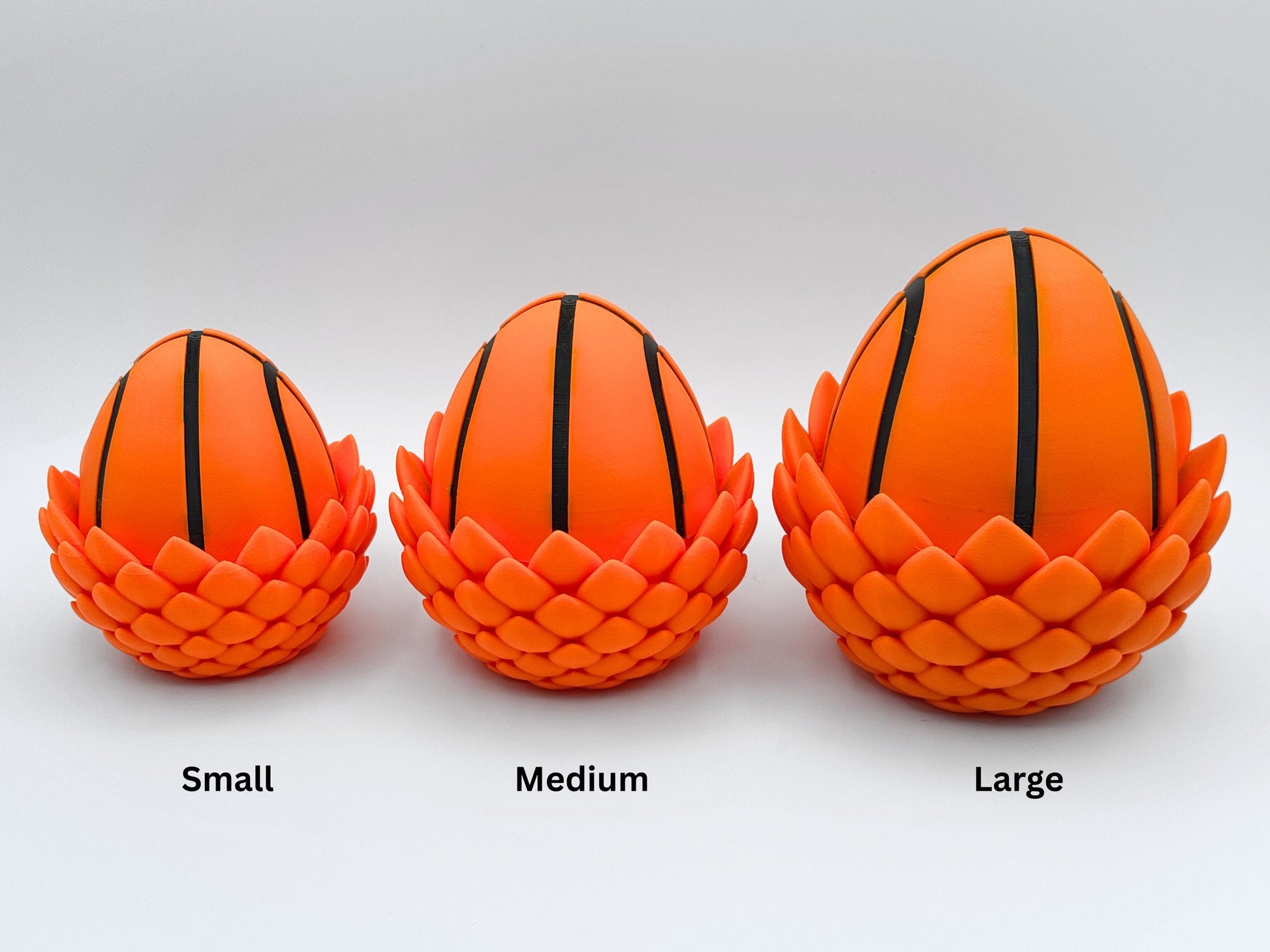 Three 3D printed dragon eggs designed to resemble basketballs, displayed in small, medium and large sizes from left to right. Each egg features bright orange panels with black lines and a textured scale like base. Ideal for use with articulated Basketball Dragon figurines as themed storage or display pieces. Shown on a clean white background with labeled sizes.