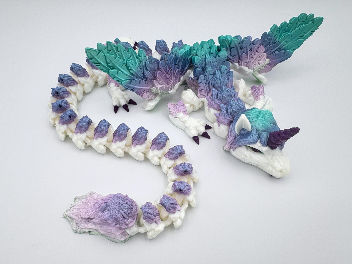 3D printed unicorn dragon lying on a flat white surface. Features a fully articulated segmented body with silk rainbow filament in teal, mint and lavender. Large feathered wings extend from its back. The dragon has a sculpted mane, deep purple unicorn horn and matching purple claws. Tail ends in a detailed plume of feathery texture. Printed with glossy finish and visible scale detailing.