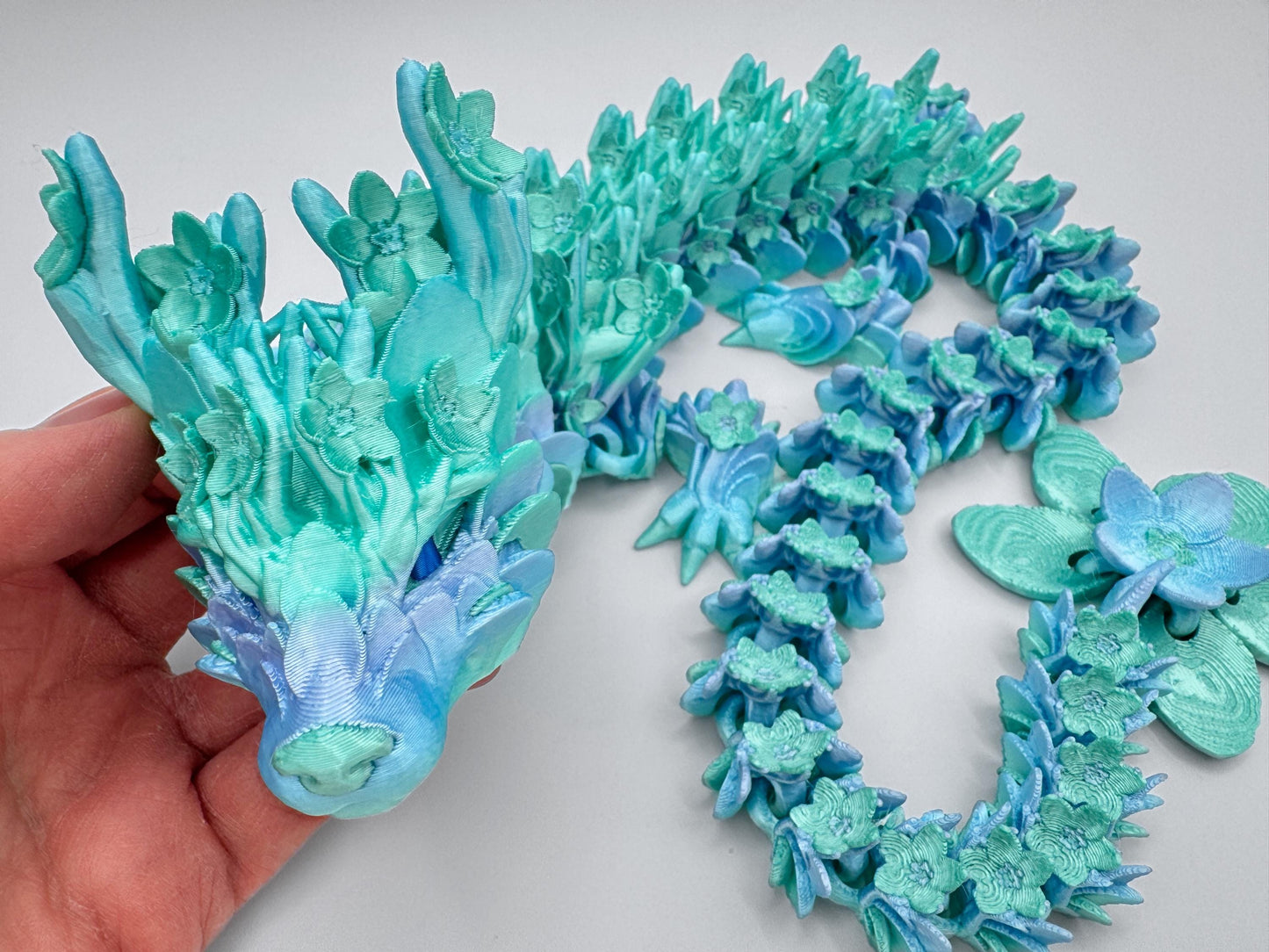 Close-up of a 3D printed articulated Cherry Blossom dragon in mint green rainbow, shown resting in a person’s hand. The dragon’s head and upper body are detailed with layered blossom accents and flowing segments, highlighting its flexible articulated design. The curled body leads into a matching blossom-shaped tail, making it a colorful floral dragon fidget or display piece.