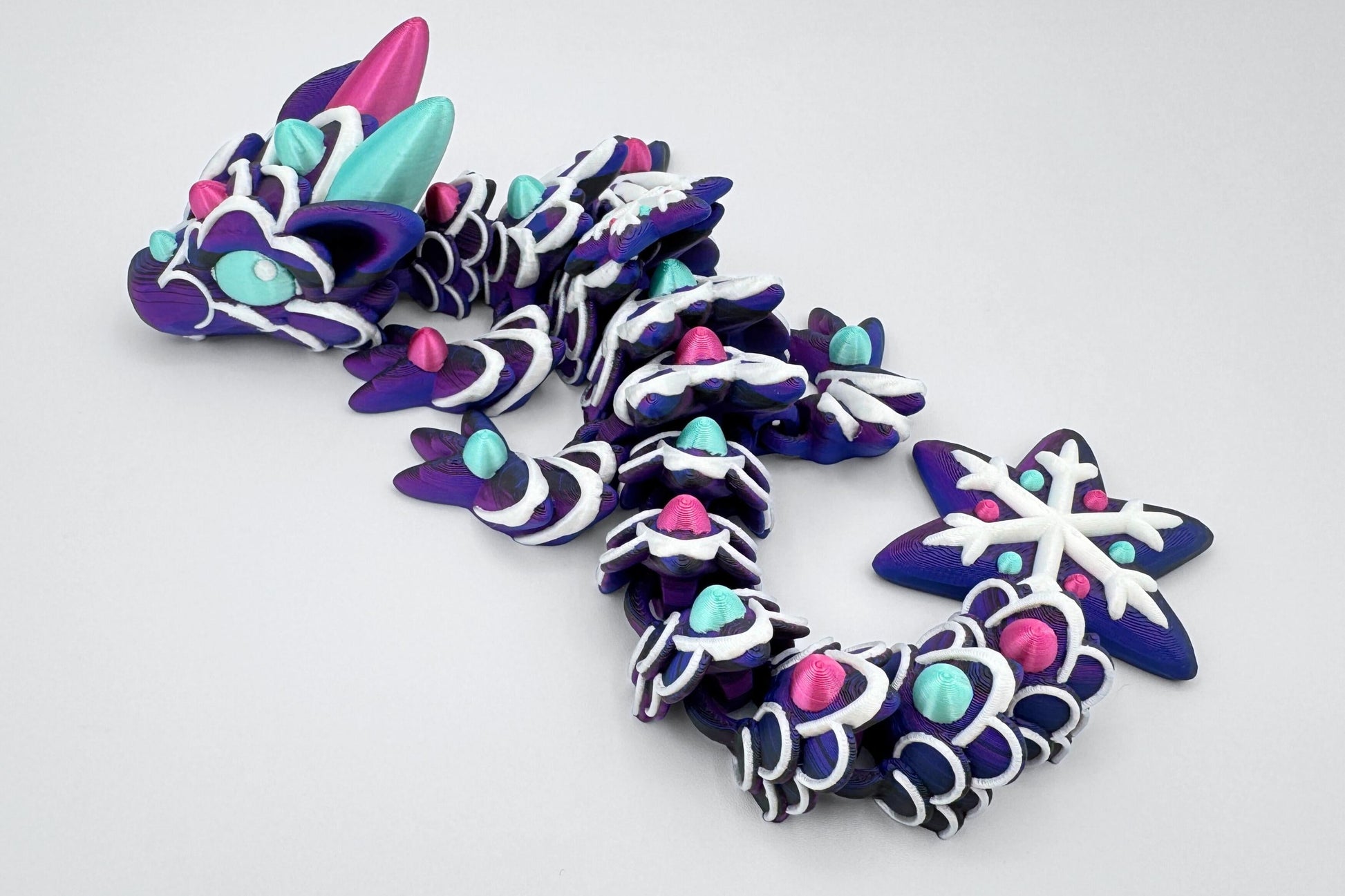 Side angle of a deep purple baby gingerbread dragon posed in a curve. Its segments are piped with white icing-like trim and topped with gumdrop-shaped accents in magenta and mint green. The dragon’s head features large candy-colored horns, ears, and eyes. The tail ends in a sculpted snowflake with white icing lines and gumdrop dots. The surface finish resembles a glossy iced cookie, flexible and colorful.