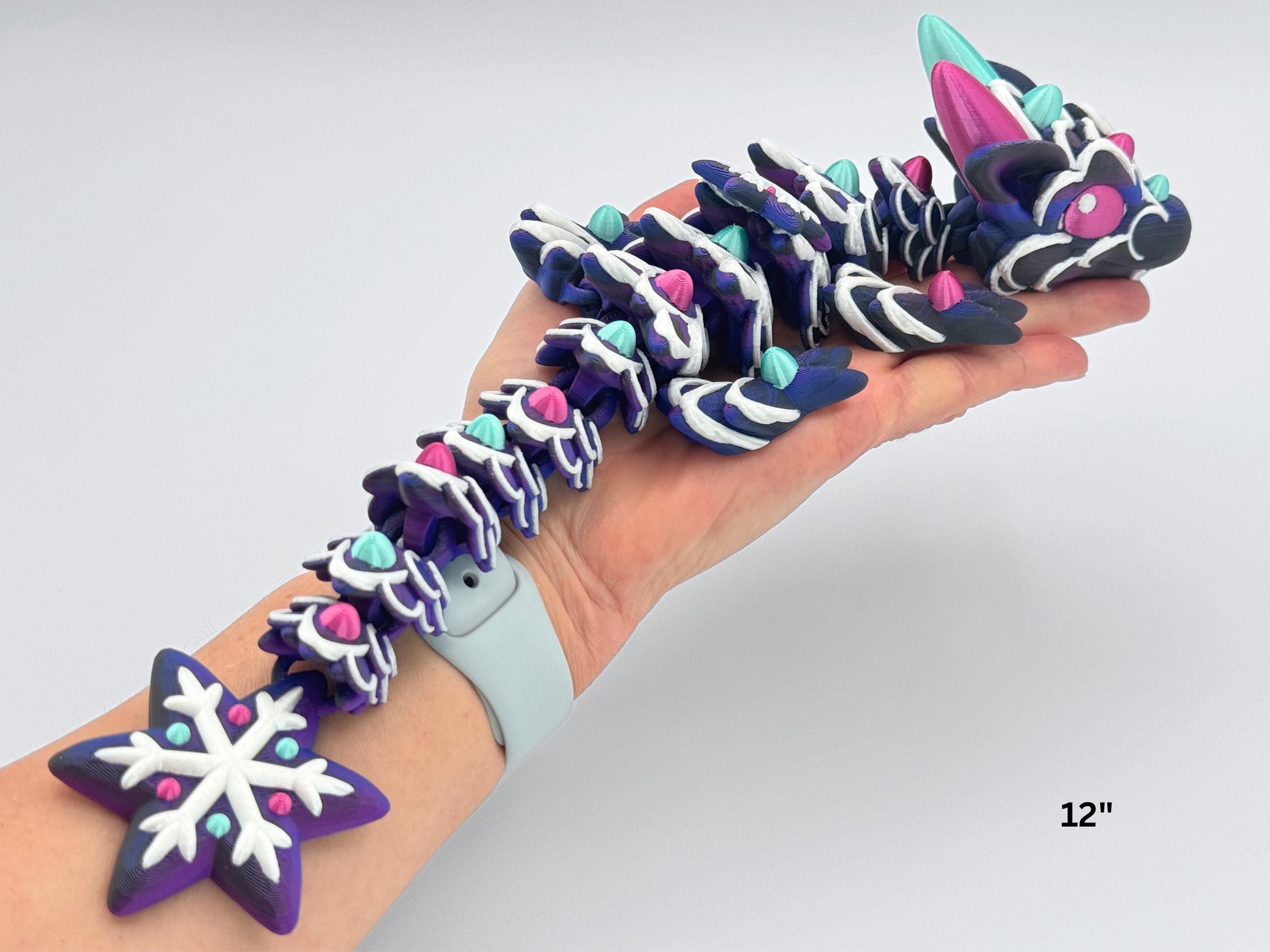 Baby gingerbread dragon shown resting in a person’s hand, about 12 inches long. Its jointed body has a purple base, decorated with white piped icing and gumdrop-like spikes in mint green and magenta. The horns and eyes match the same bright candy theme. The tail ends in a detailed six-point snowflake with icing and gumdrop dots. Each segment moves and reflects a holiday cookie theme.