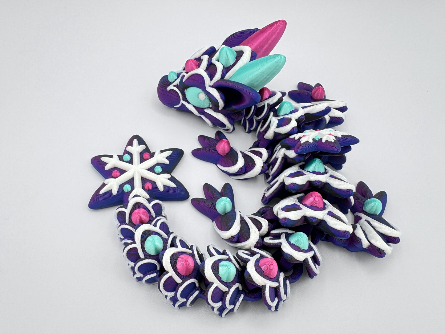 Top-down view of baby gingerbread dragon arranged in a spiral. The body is printed in a dark purple tone, with white icing-style piping and alternating magenta and mint green gumdrop-like accents. The face has two large horns and candy-decorated eyes. Each limb is decorated with icing, and the tail ends in a star-shaped snowflake with raised gumdrop dots. The full piece resembles a sculpted holiday treat.