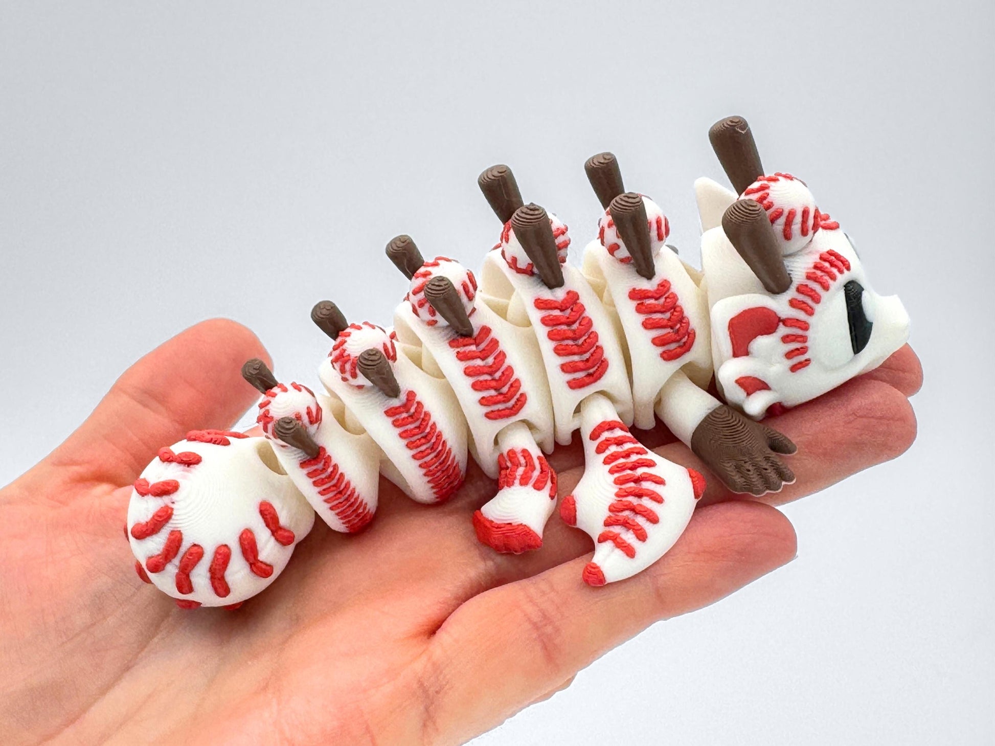 One Baseball Dragon figurine curled slightly in a person’s hand for size reference. The articulated white body features red baseball stitching, brown bat-like spines, and black expressive eyes.