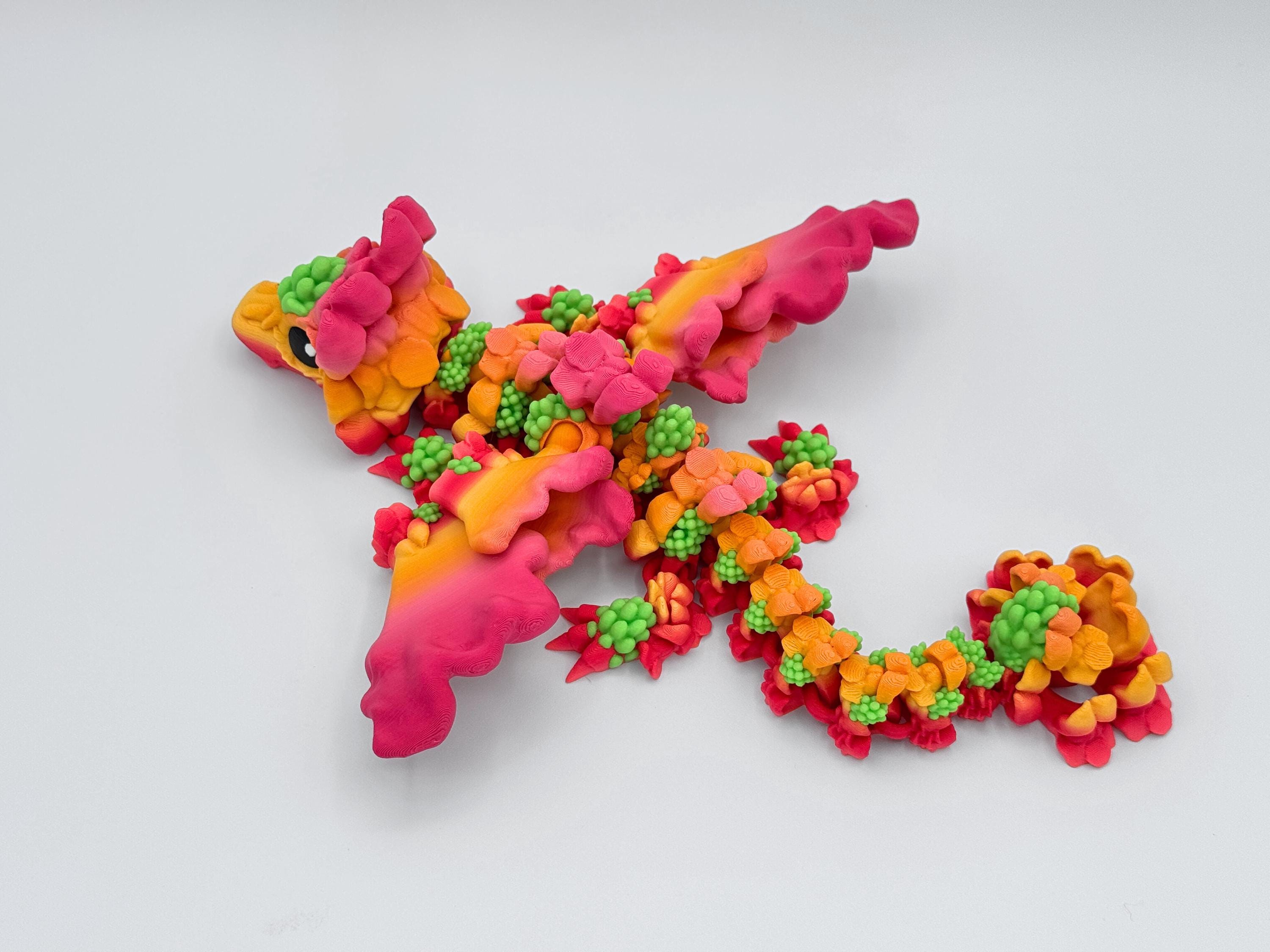 Top-down view of a Snapdragon Dragon figurine in a flight pose with wings extended. The body is curved with detailed flower-like segments in yellow, pink, orange, and green. The head is turned slightly to the side. Background is clean and white.