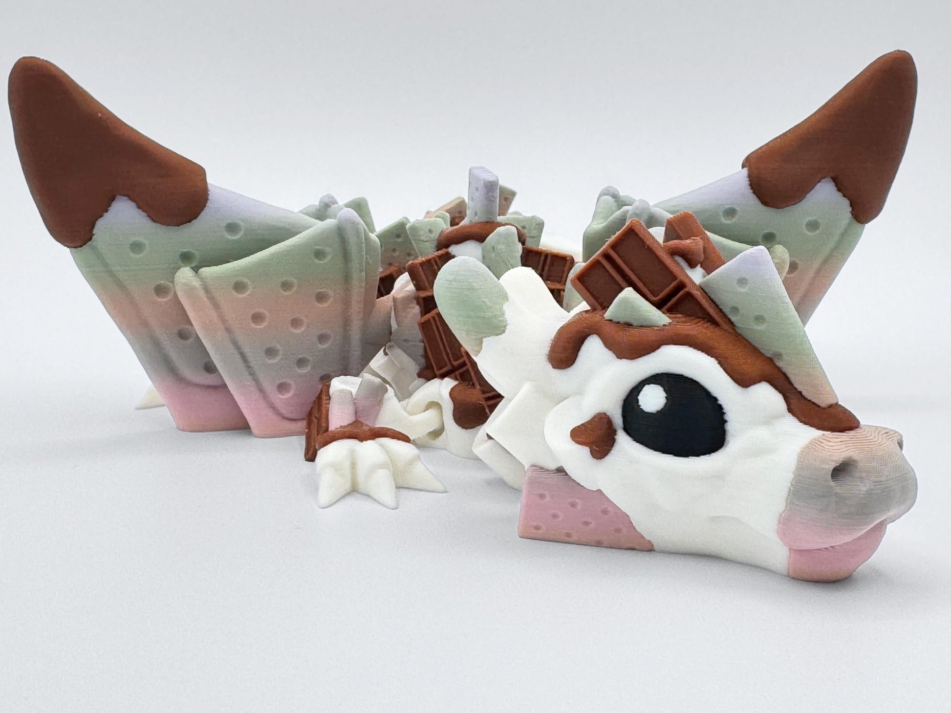 Close-up frontal view of the S’mores Dragon figurine, showing wide eyes, chocolate-graham and creamy marshmallow facial textures. The cracker wings rise behind the head with gradient color shifts from green to pink. Chocolate bar clusters are placed along the neck. Designed as a dessert-themed articulated fidget dragon, this 3D printed collectible blends soft candy hues with fantasy styling for display or sensory play.