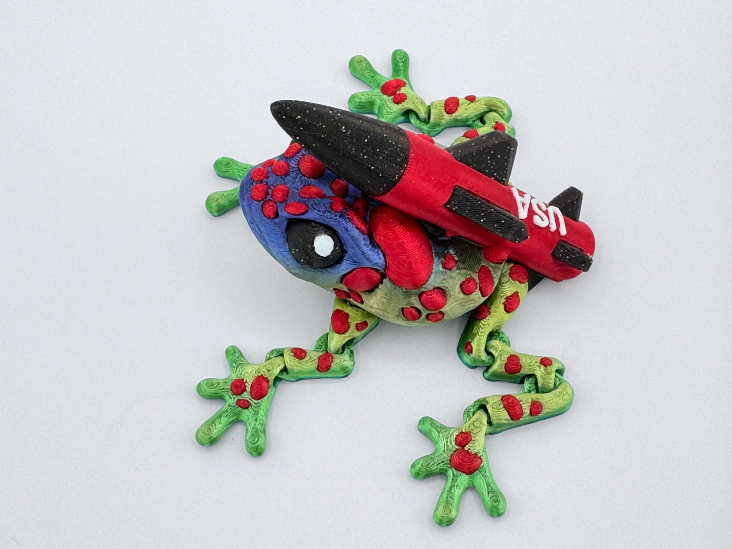 3D printed Missile Toad frog in festive colors, featuring a green-to-blue color-shift body with raised red spots and a bright red-and-black rocket labeled "USA" mounted on its back. Detailed textured skin, large expressive eyes, and poseable legs bring playful character. The red and green palette gives it a fun Christmas-themed look with a bold twist.
