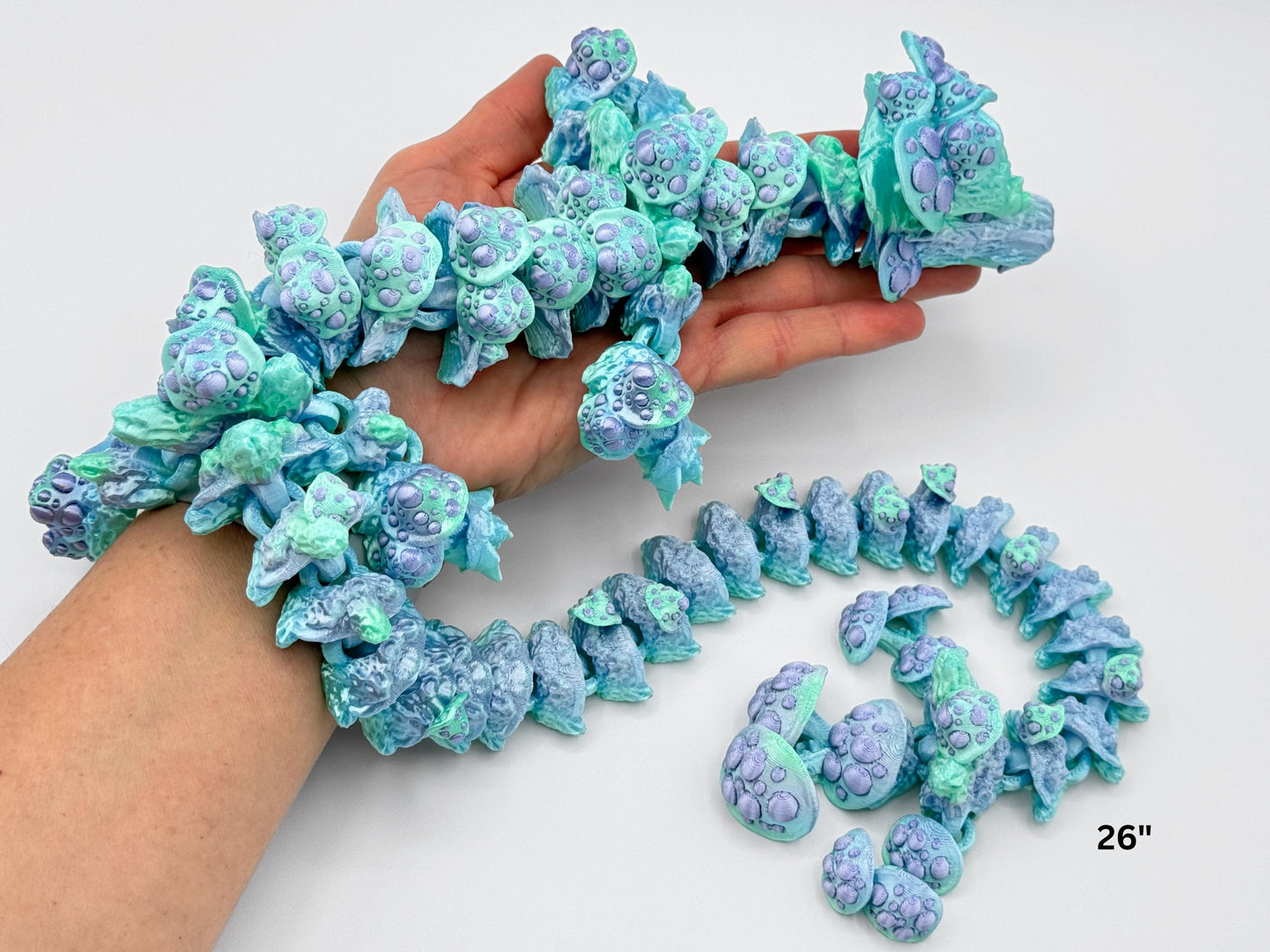 Hand holding the mushroom dragon, showing its full articulated body draped over the wrist. The figurine blends icy mint, pale aqua, and lavender hues with delicate mushroom caps on each flexible segment, highlighting its size and movement.