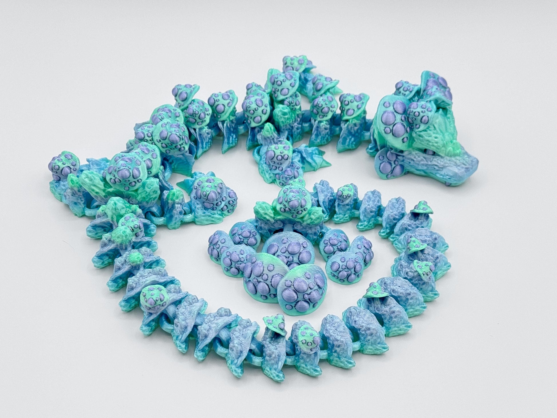 3D printed mushroom dragon curled in a loose spiral, showing off pastel green and blue body segments with soft lavender mushroom caps. The articulated design highlights smooth ridges and textured mushroom details running down the spine and tail.