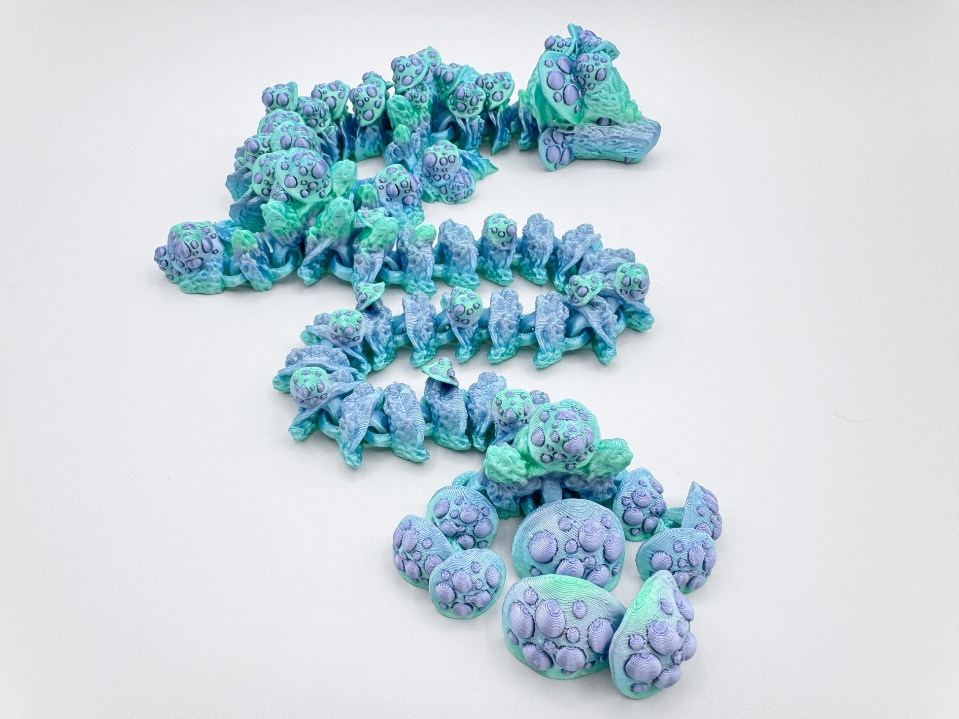 Top-down view of a pastel green and lavender mushroom dragon. The articulated segments trail down in a gentle curve, each one featuring textured mushrooms in soft purples on a marbled aqua-blue base. The dragon’s head is visible at the top.