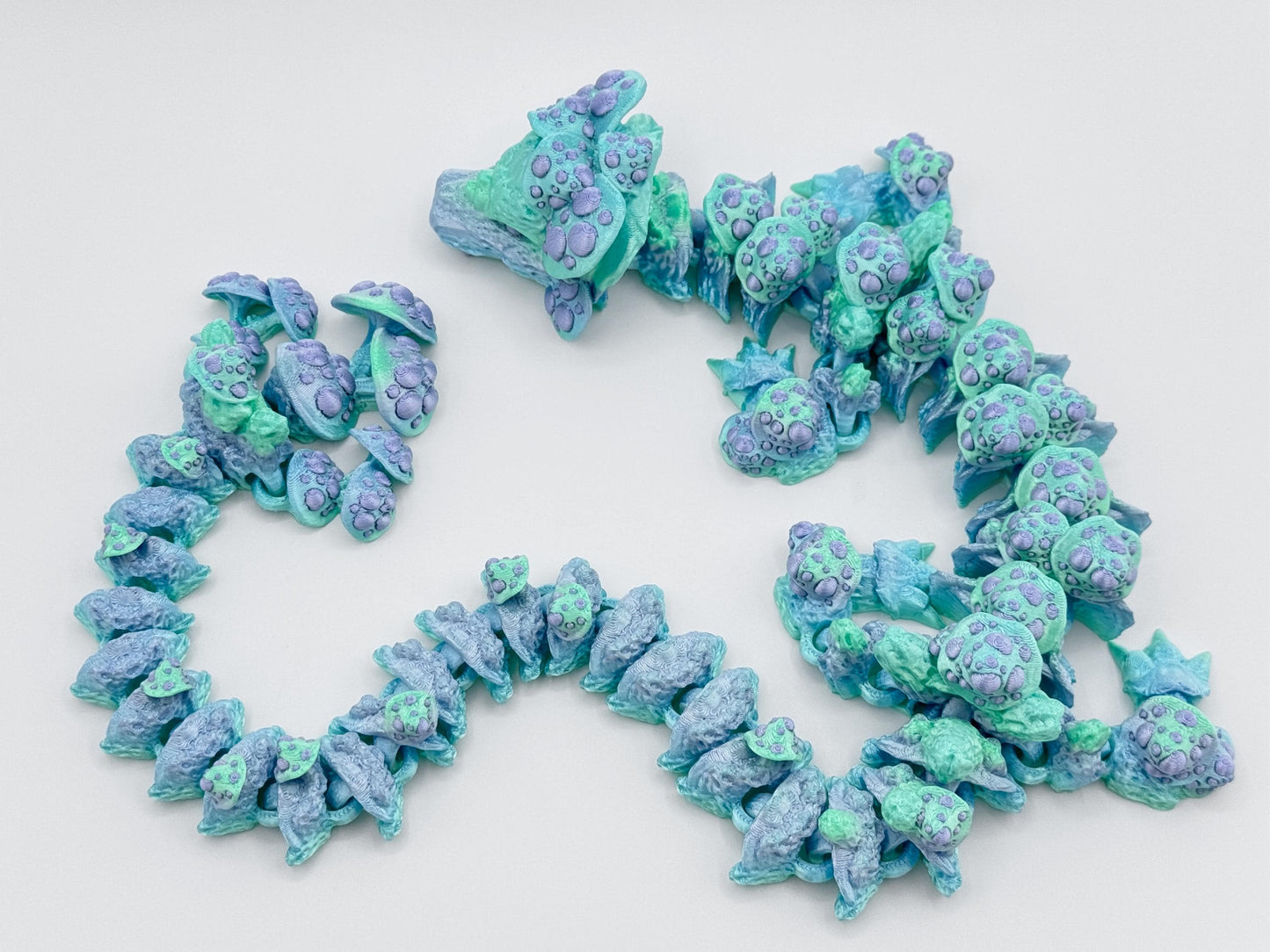 Close-up angle of the mushroom dragon curled in an open spiral. The dragon’s pastel green head is covered in lavender mushrooms, with a flowing body of articulated blue and aqua segments. Light texturing adds a mossy, magical vibe.