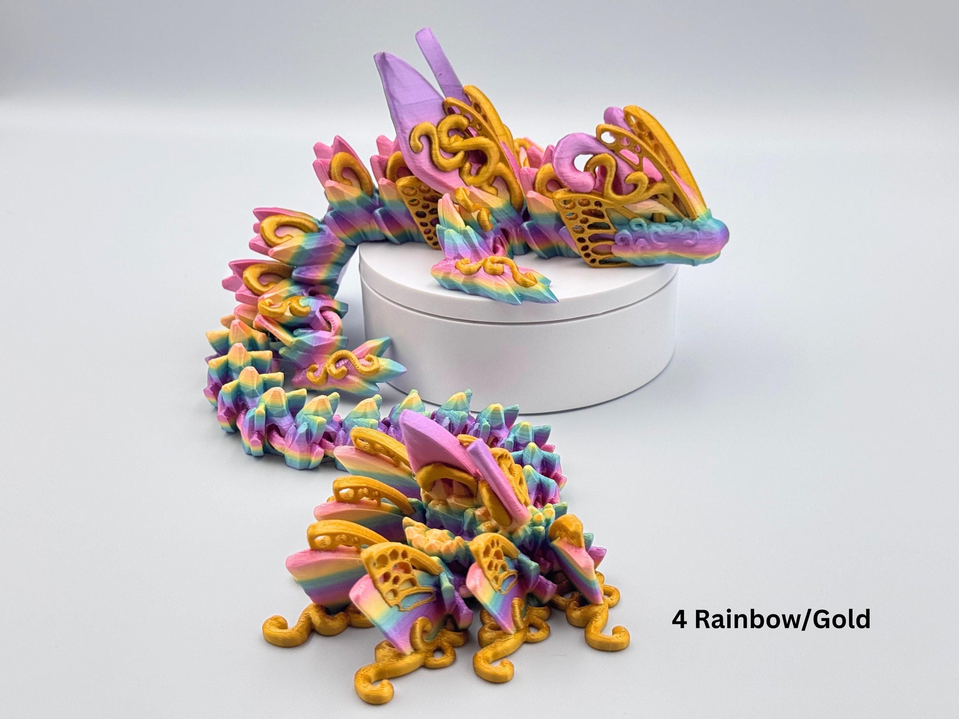 3D printed butterfly dragon figurine in rainbow filament with gold accents and details, shown curled on a white stand with articulated segments and sculpted texture.