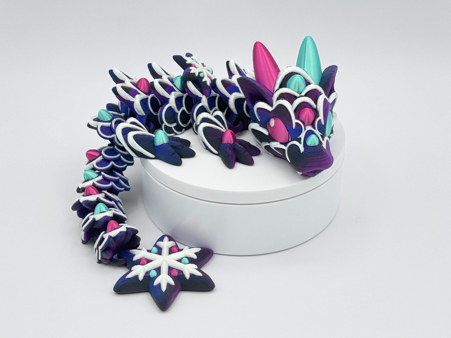 3D Printed Gingerbread dragon posed in a curled shape on a white display stand. Its body is a rich purple gradient with white icing-style piping on each segment. Mint green and magenta gumdrop-like accents sit along the back, limbs, and head. The dragon has large pointed horns and wide eyes, giving it a bright, animated look. The tail ends in a snowflake shape with frosted white icing and matching gumdrop details.