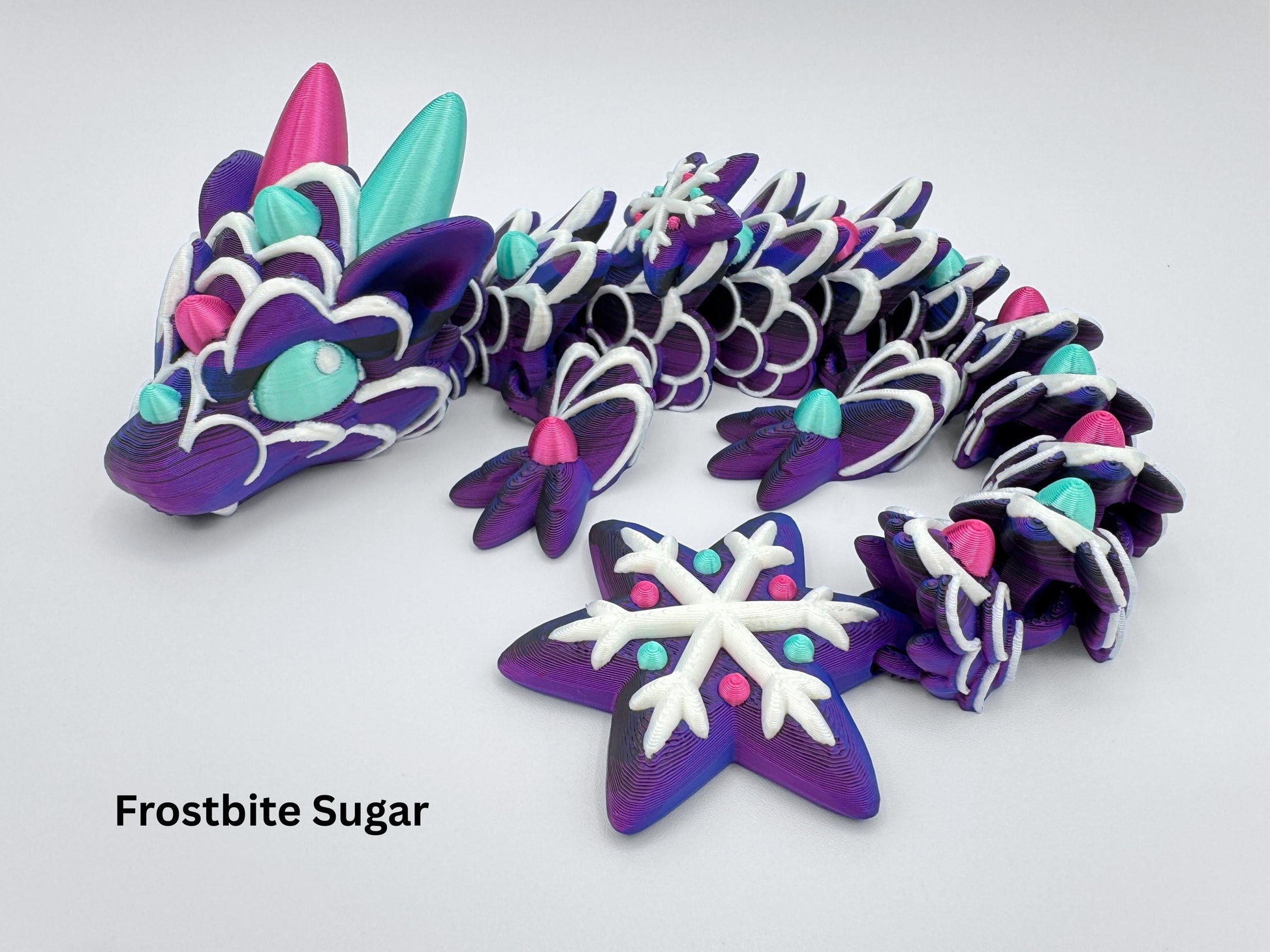 Articulated gingerbread dragon in a side pose with label “Frostbite Sugar.” The figure features a purple cookie-like body with white icing lines and evenly spaced gumdrop decorations in magenta and mint green. Horns and claws are sculpted in the same candy color palette. A snowflake-shaped tail with raised white icing and gumdrop dots completes the festive design. Segments are rounded and moveable.