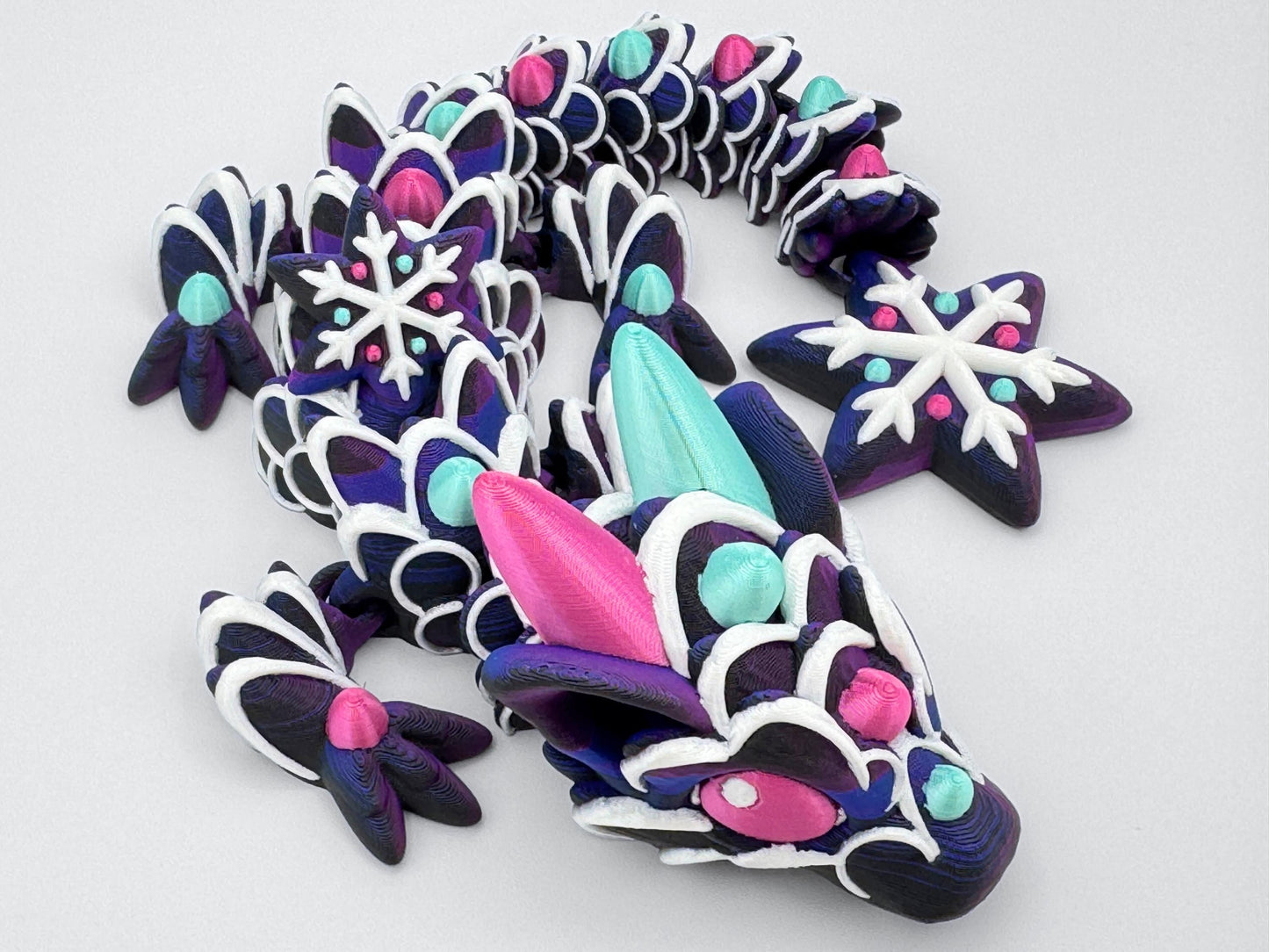 Close-up of the baby gingerbread dragon’s face and upper body. The head is detailed with swirled white icing lines, two large horns in mint green and magenta, and candy-like eyes. Gumdrop accents continue down the back and limbs. The tail features a six-pointed snowflake decorated with piped icing and colorful dots. The dragon is printed in a dark purple gradient, giving the look of a frosted cookie fidget.