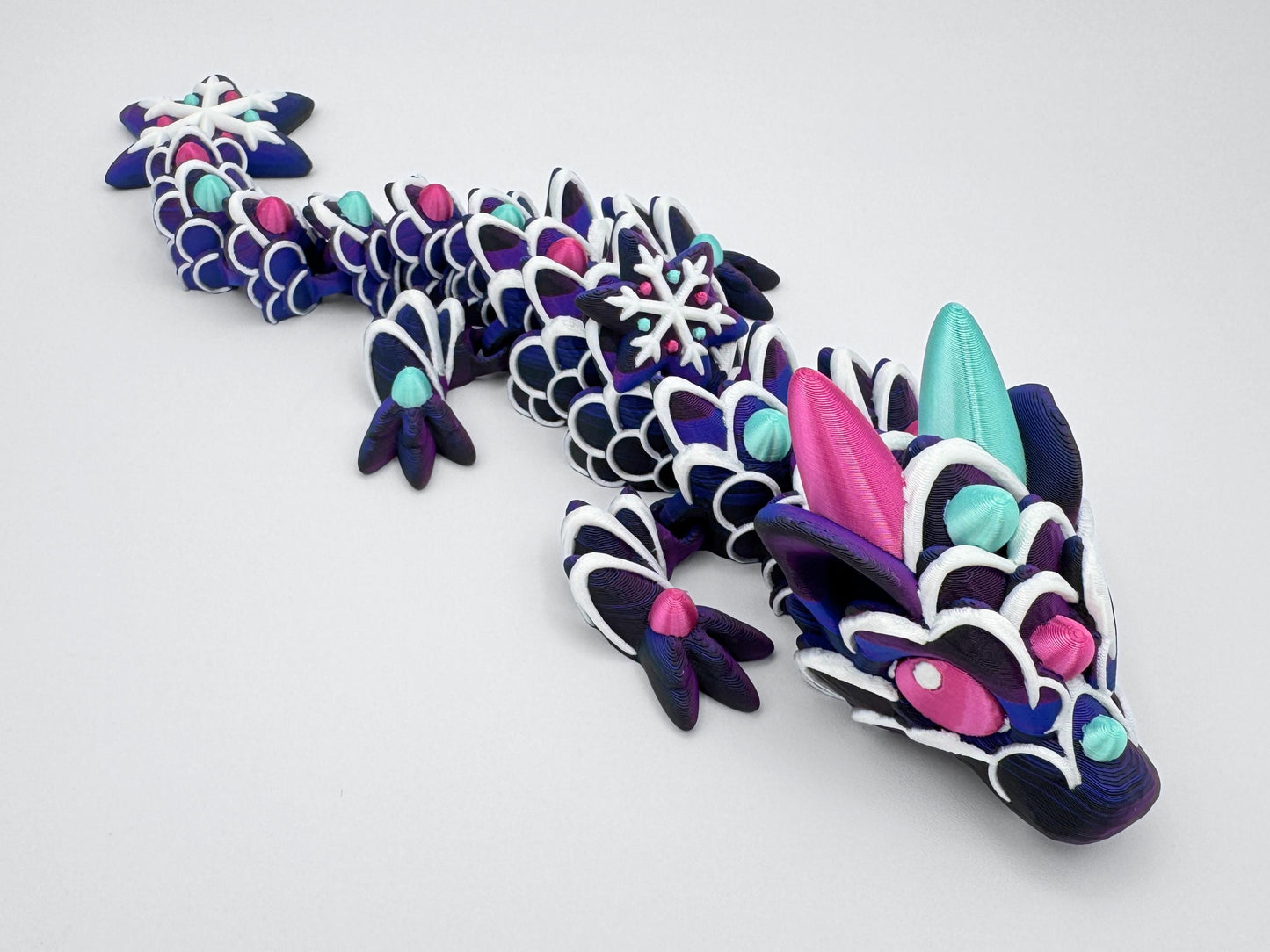 Full view of the baby gingerbread dragon stretched across a light background. The figure has a dark purple cookie-like base with white piped icing and raised gumdrop accents in alternating mint green and magenta. Large horns, ears, and detailed eyes give the head an expressive look. The tail ends in a snowflake-shaped segment with white icing and gumdrop-style decorations to match the rest of the body.