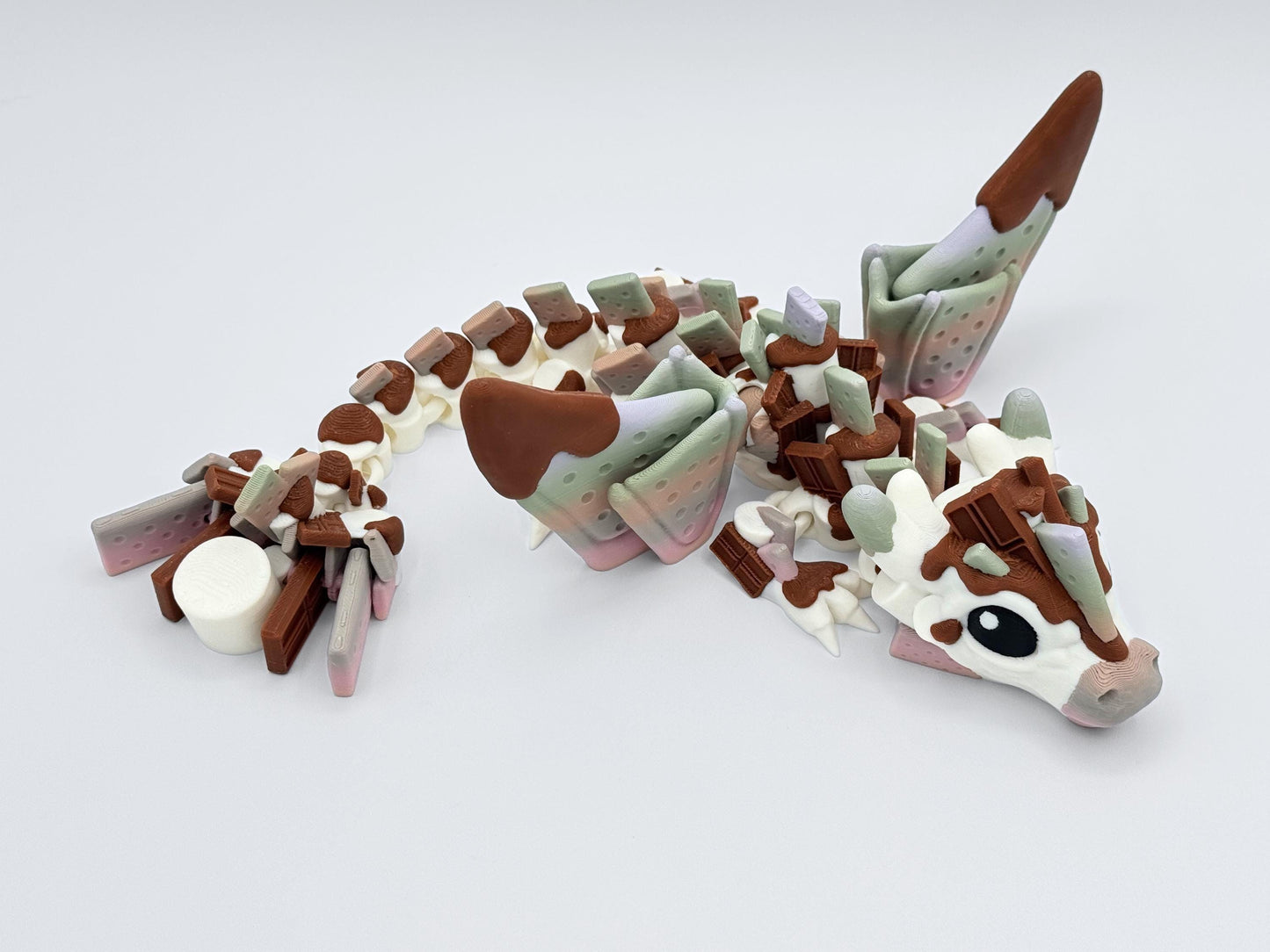 3D printed S’mores Dragon figurine shown from the top, fully extended on a glossy surface. Wings feature perforated graham cracker panels dipped in chocolate, with pastel pink and mint highlights. Body includes marshmallow-textured joints and chocolate bar segments along the spine. This fantasy fidget collectible is styled to resemble a dessert dragon with rich details and smooth articulation, great for display or as a dessert-themed dragon gift.