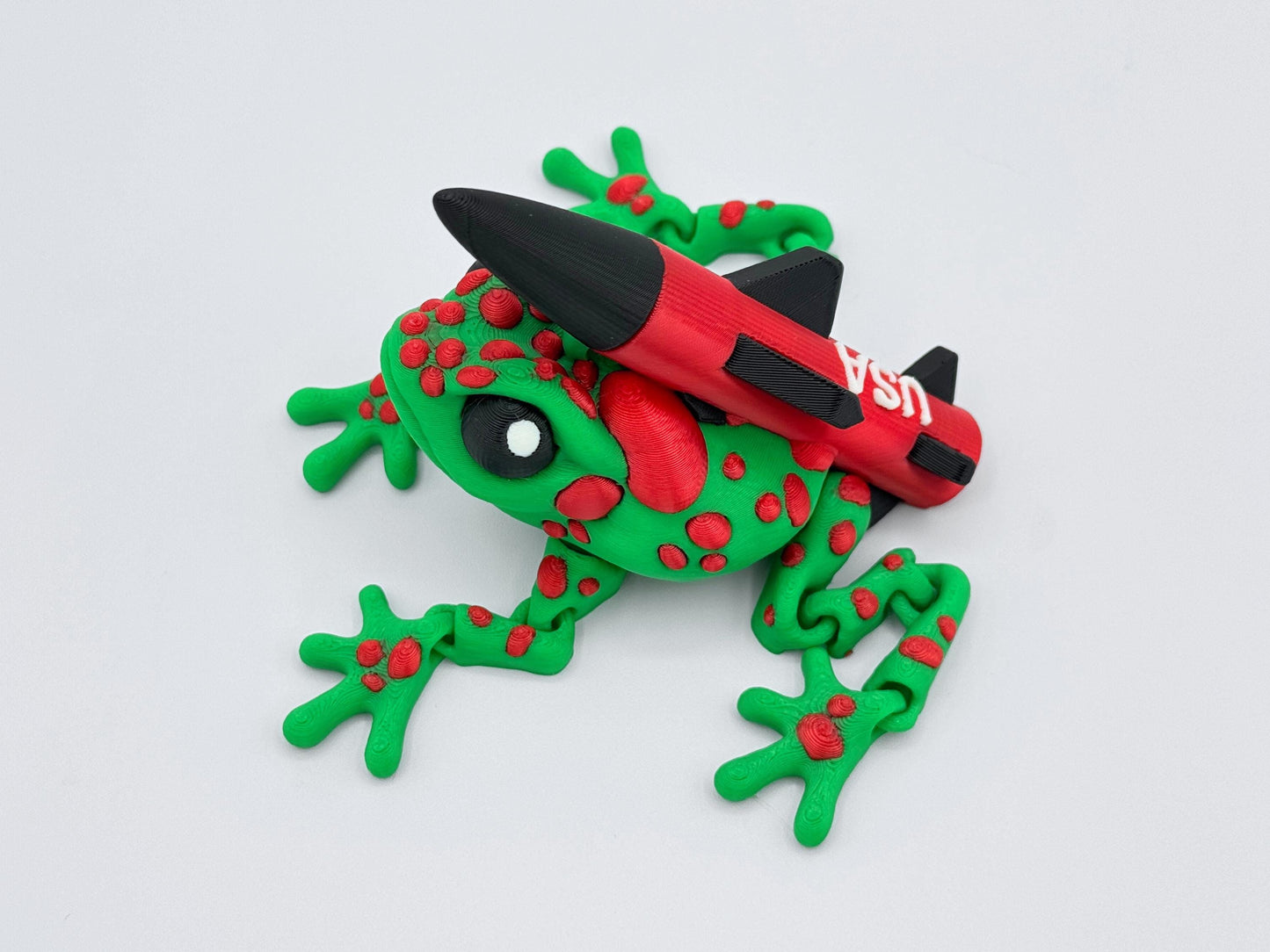 Overhead view of the articulated Missile Toad — a green frog covered in raised red spots and equipped with a bold red and black rocket on its back. The rocket features black fins and the word “USA” in white lettering.