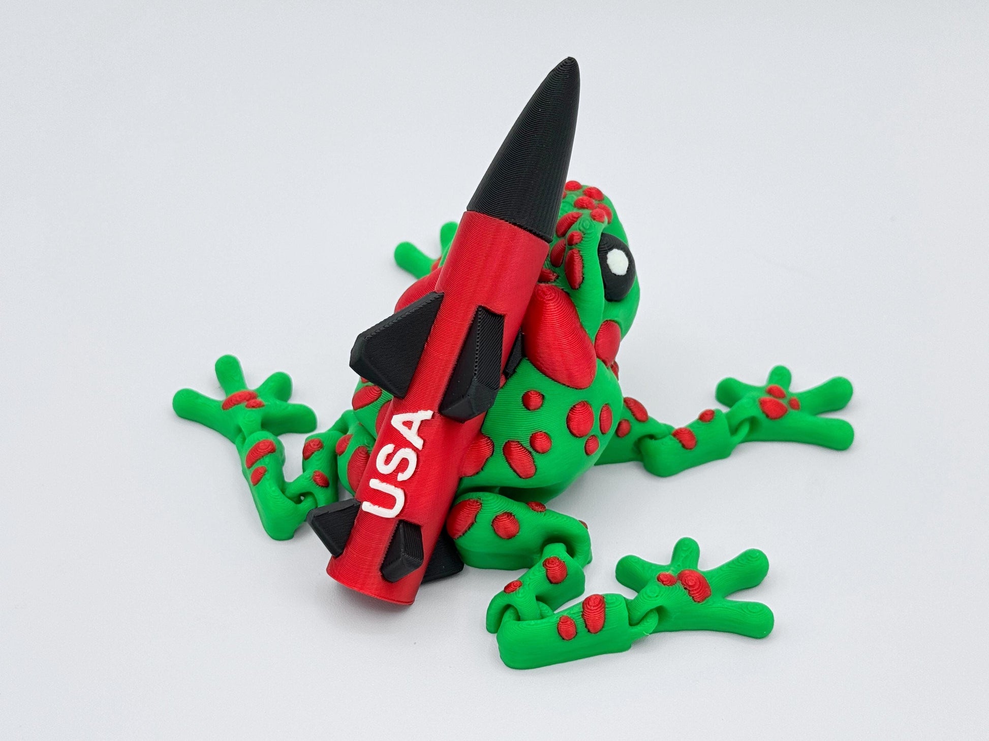 Back view of the 3D printed Missile Toad figurine showing full rocket detail, including all four fins, engraved "USA" text, and the frog’s leg articulation. The red spots trail from the head down its spine and limbs.