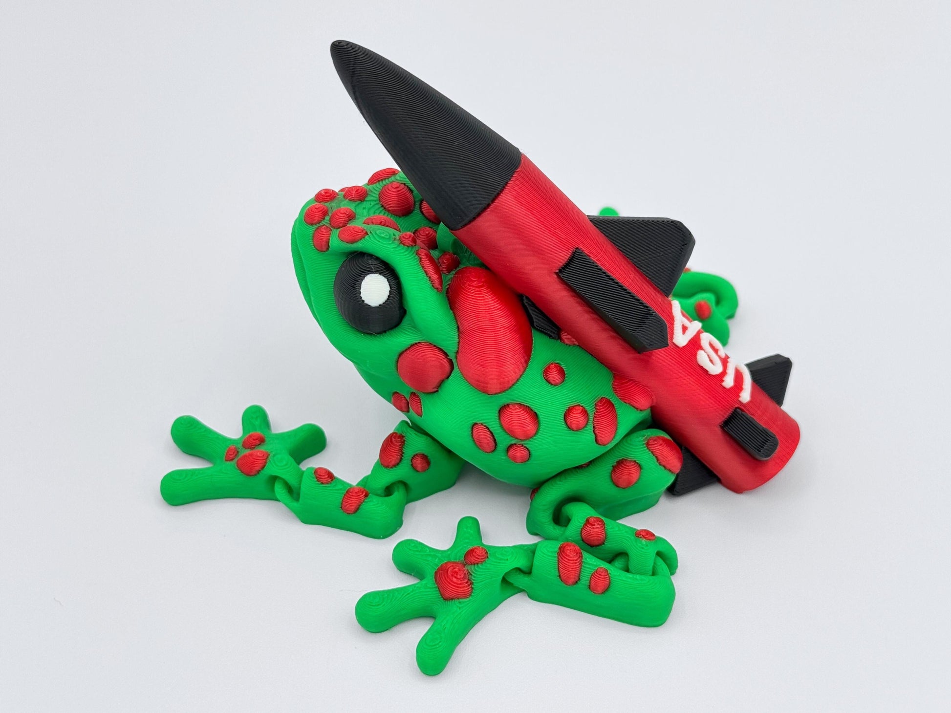 Green and red 3D printed frog figurine with a large red and black rocket labeled "USA" mounted on its back. The frog’s pose and expressive eyes add character, with sculpted red bumps across its vibrant green body for a cartoon like appearance.