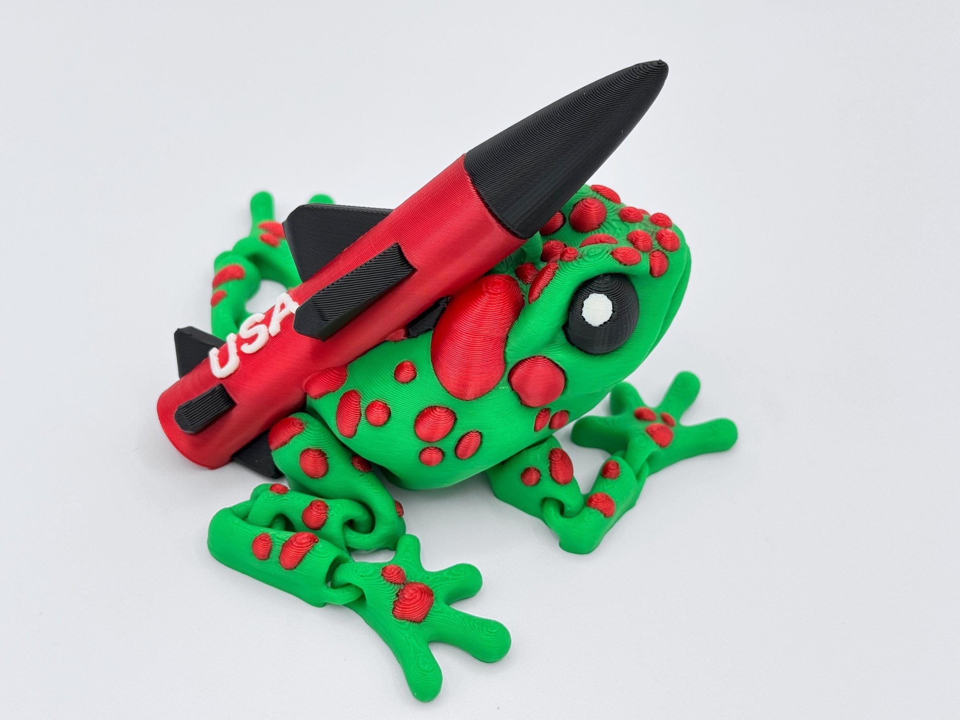 Side profile of the Missile Toad figurine showing off its large black and white cartoon-style eye, red textured spots, and detailed missile fins. The frog's colorful body curves naturally in a crouched stance.
