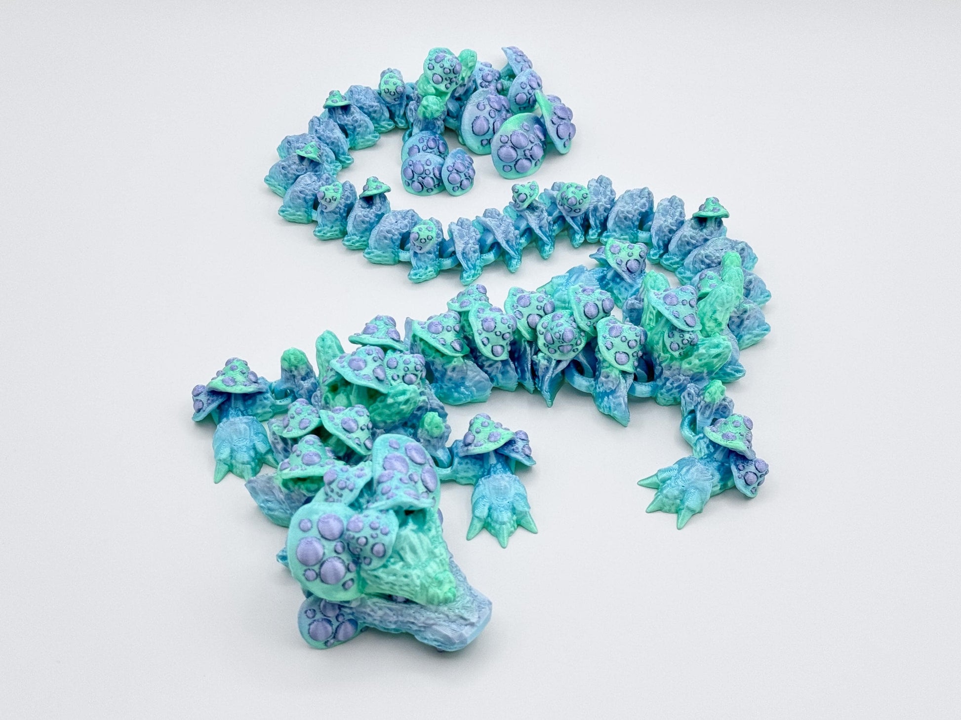 Side angle of the 3D printed mushroom dragon with its body coiled in a reverse-S shape. Pale mint green, aqua and soft periwinkle hues fade along the dragon’s limbs and mushrooms, emphasizing its articulated build and fantasy woodland style.