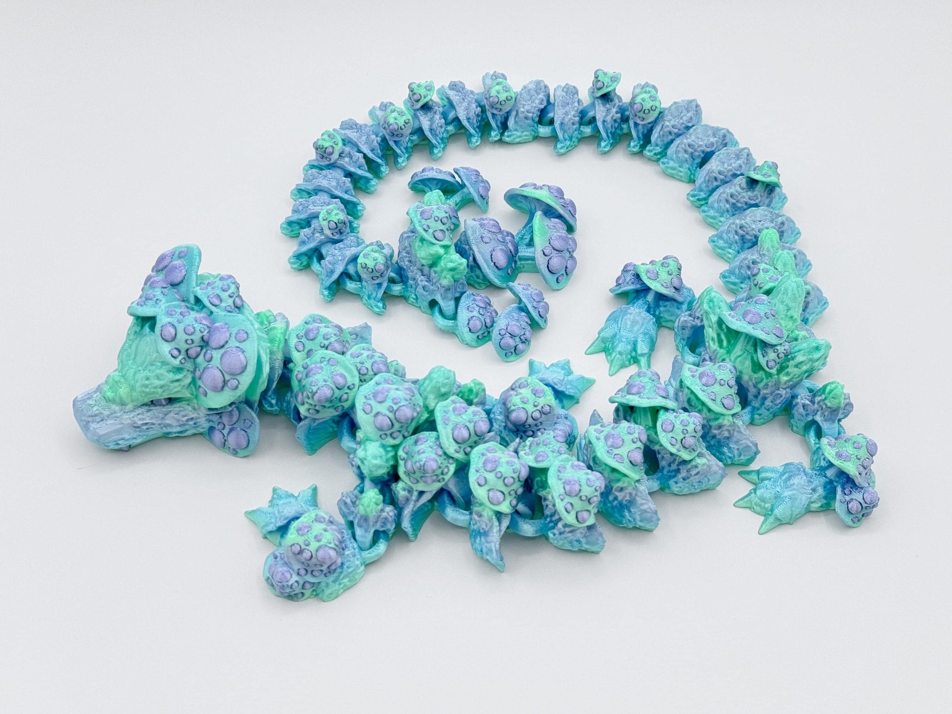 Full view of the 3D printed articulated mushroom dragon positioned in a rounded spiral. The dragon’s body fades from icy mint to pale blue, with soft purple mushroom textures scattered across its back, tail, and face for a gentle fantasy aesthetic.