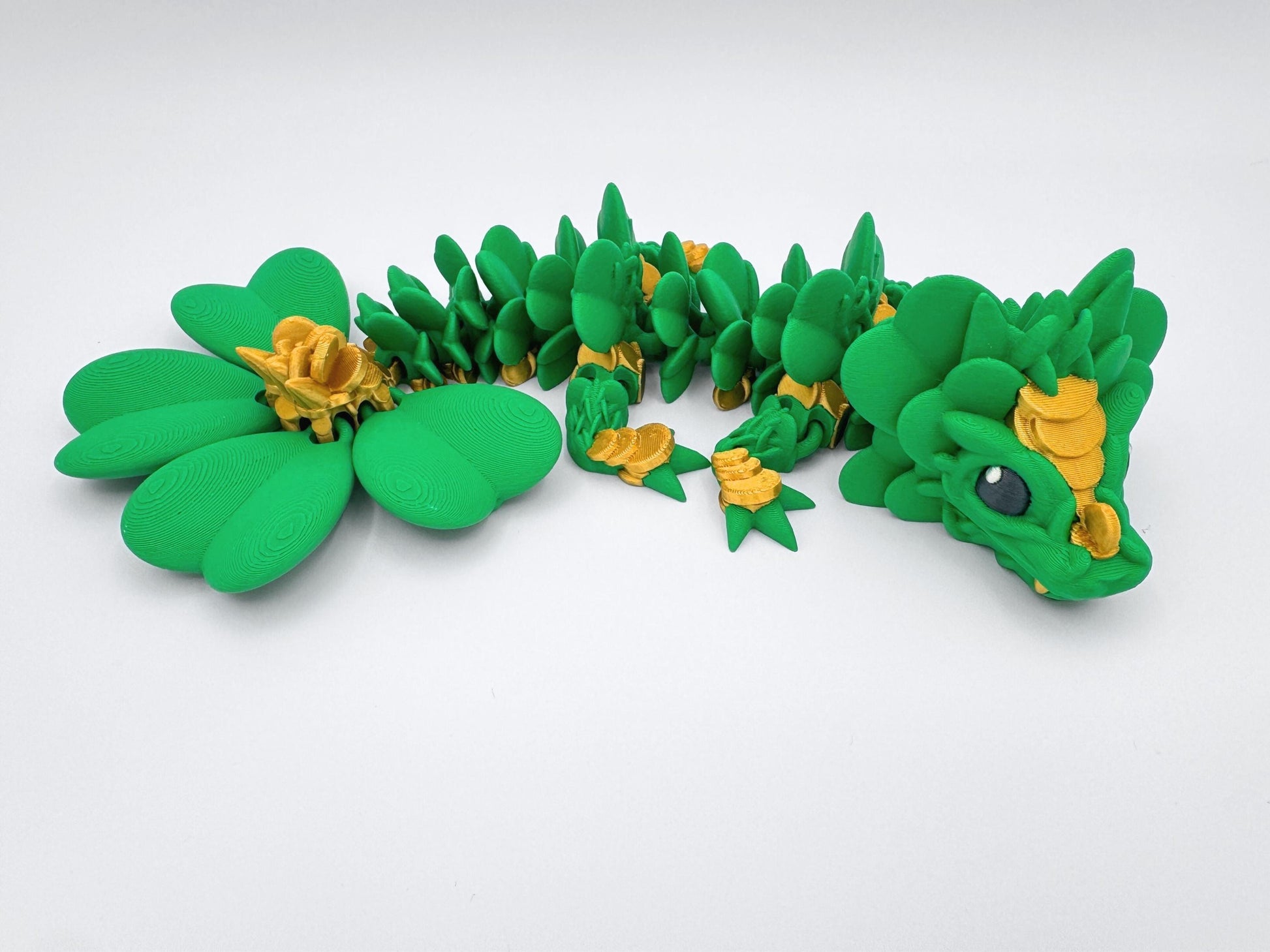 3D Printed Clover Dragon green gold and black articulated figurine with a clover 4 leaf design along its body. The dragon is made up of many small, overlapping leaves and has a large head with a wide mouth and large eyes. The dragon's tail is curled up behind it and is lying on a white surface.  Used as desk decor for seasonal gift for St. Patrick’s Day.