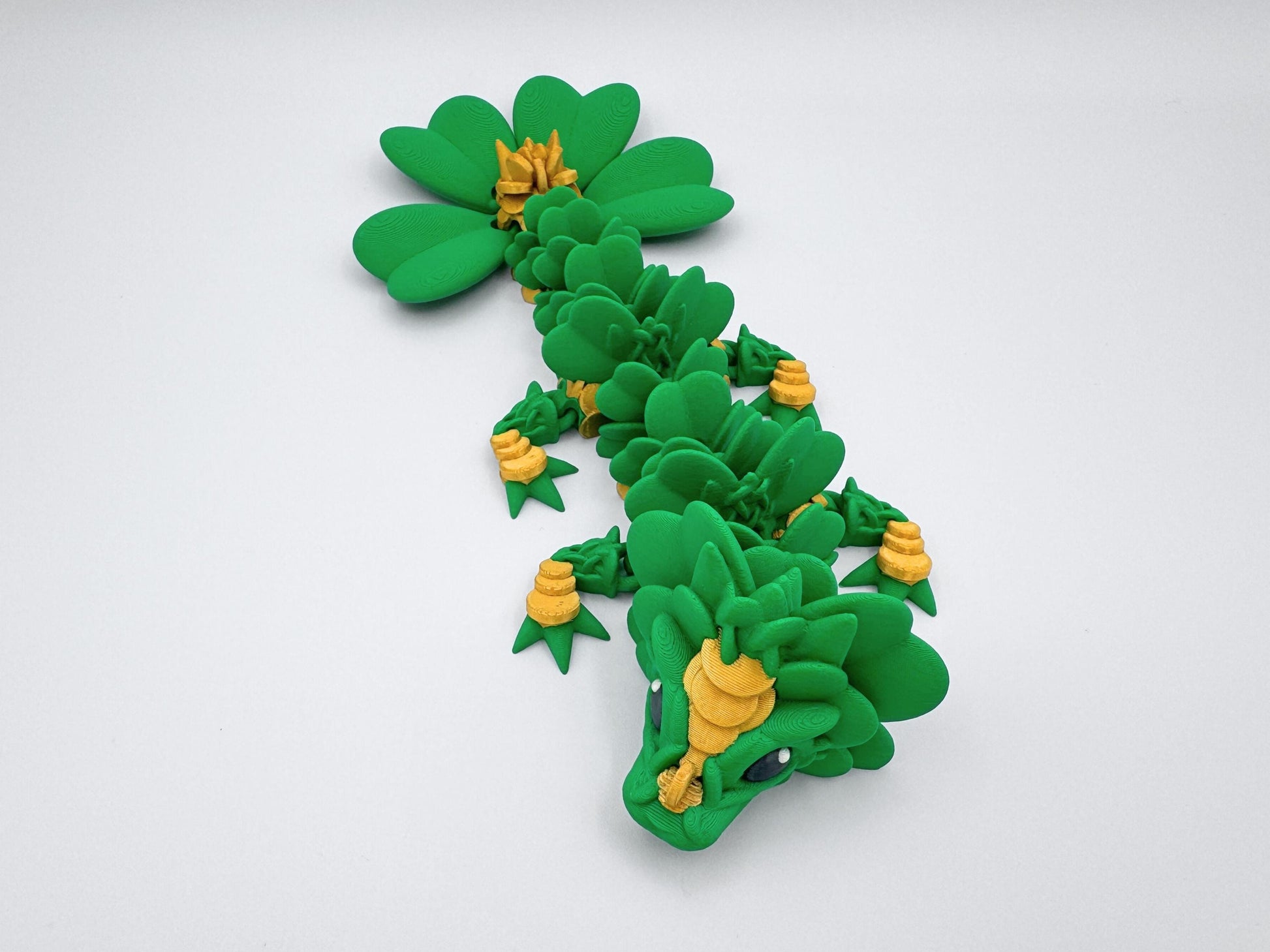 3D Printed Clover Dragon green gold and black articulated figurine with a clover 4 leaf design along its body. The dragon is made up of many small, overlapping leaves and has a large head with a wide mouth and large eyes. The dragon's tail is curled up behind it and is lying on a white surface.  Used as desk decor for seasonal gift for St. Patrick’s Day.