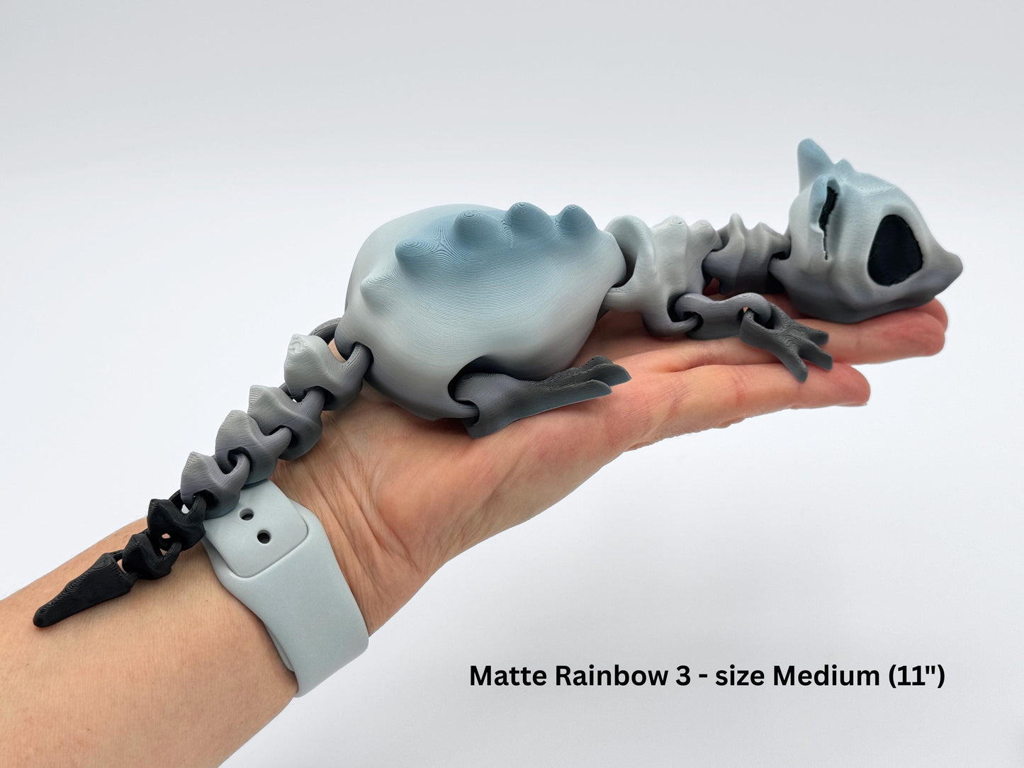 3D printed articulated bone rat figurine, 11 inches long, resting on a hand for scale. Printed in gradient gray filament with a black tail tip, segmented spine, and jointed limbs. Large black eye sockets give it a skeletal look. Smooth joints allow posing of head, legs, and tail.