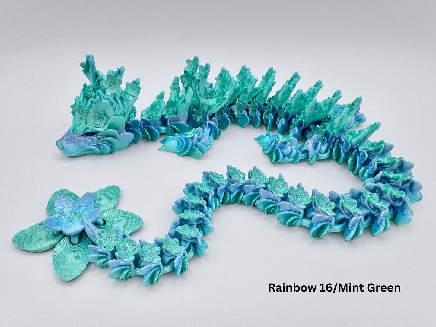 A 3D printed articulated Cherry Blossom dragon in mint green rainbow silk color, with flower like details along its head, back and tail. The dragon is stretched in a gentle S-curve, showing the flexible segmented body and layered floral textures. A large blossom shaped tail piece completes the look, making it a colorful floral dragon fidget or display figure.