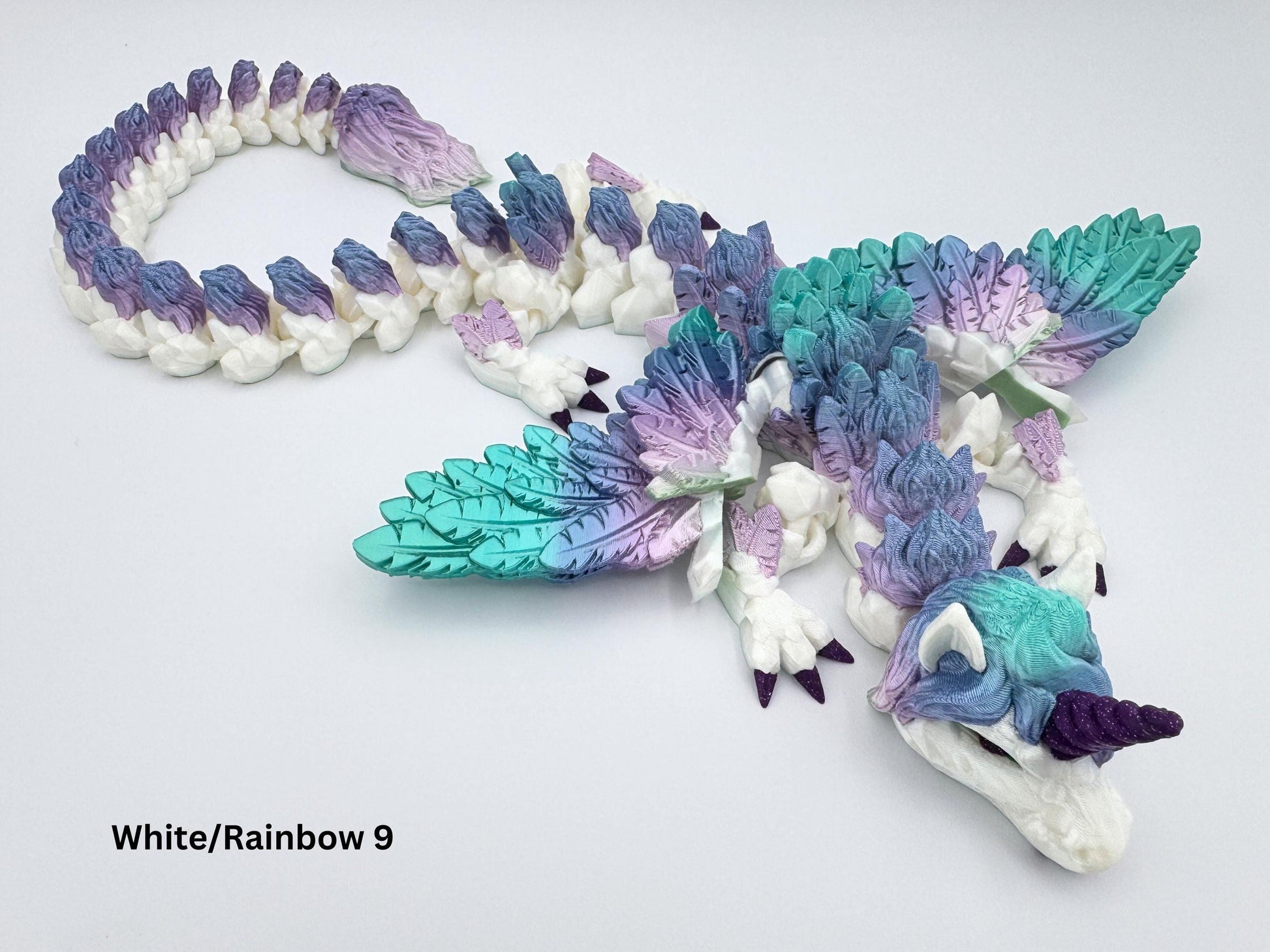 3D printed unicorn dragon lying in a loose spiral on a flat white surface. Features a fully articulated segmented body with silk rainbow filament in teal, mint and lavender. Large feathered wings extend from its back. The dragon has deep purple unicorn horn and matching purple claws. Tail ends in a detailed plume of feathery texture. Printed with glossy finish and visible scale detailing.