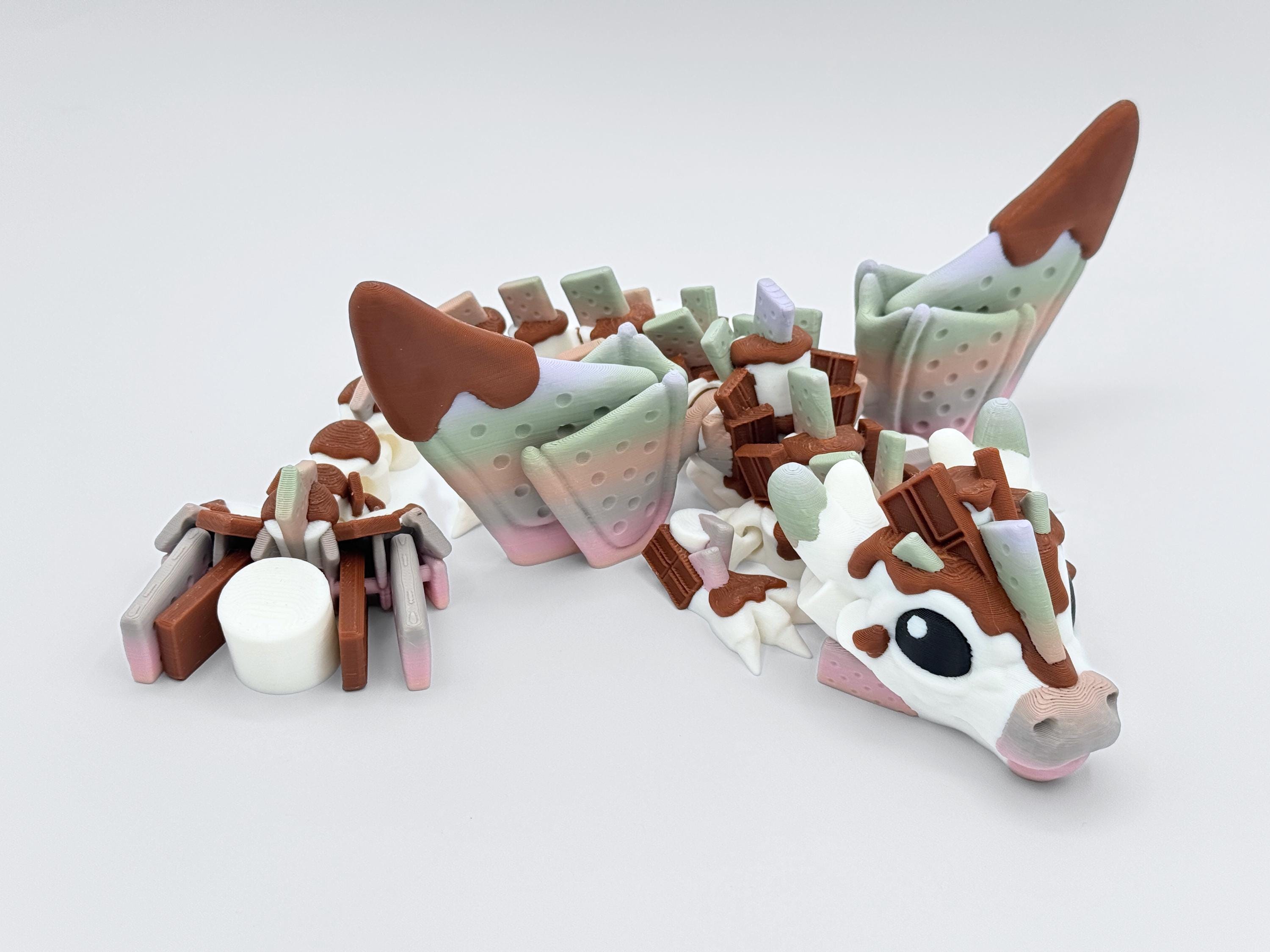 Articulated S’mores Dragon lying in a curved pose with head and tail angled forward. Dragon has chocolate-dipped cracker wings and candy-inspired spikes along its back, blending white, brown, pink and green. Its fidget-style body bends gently, and a large marshmallow detail anchors the tail. This fantasy dessert dragon is 3D printed and styled as a whimsical graham cracker collectible for dragon lovers or dessert-themed decor fans.