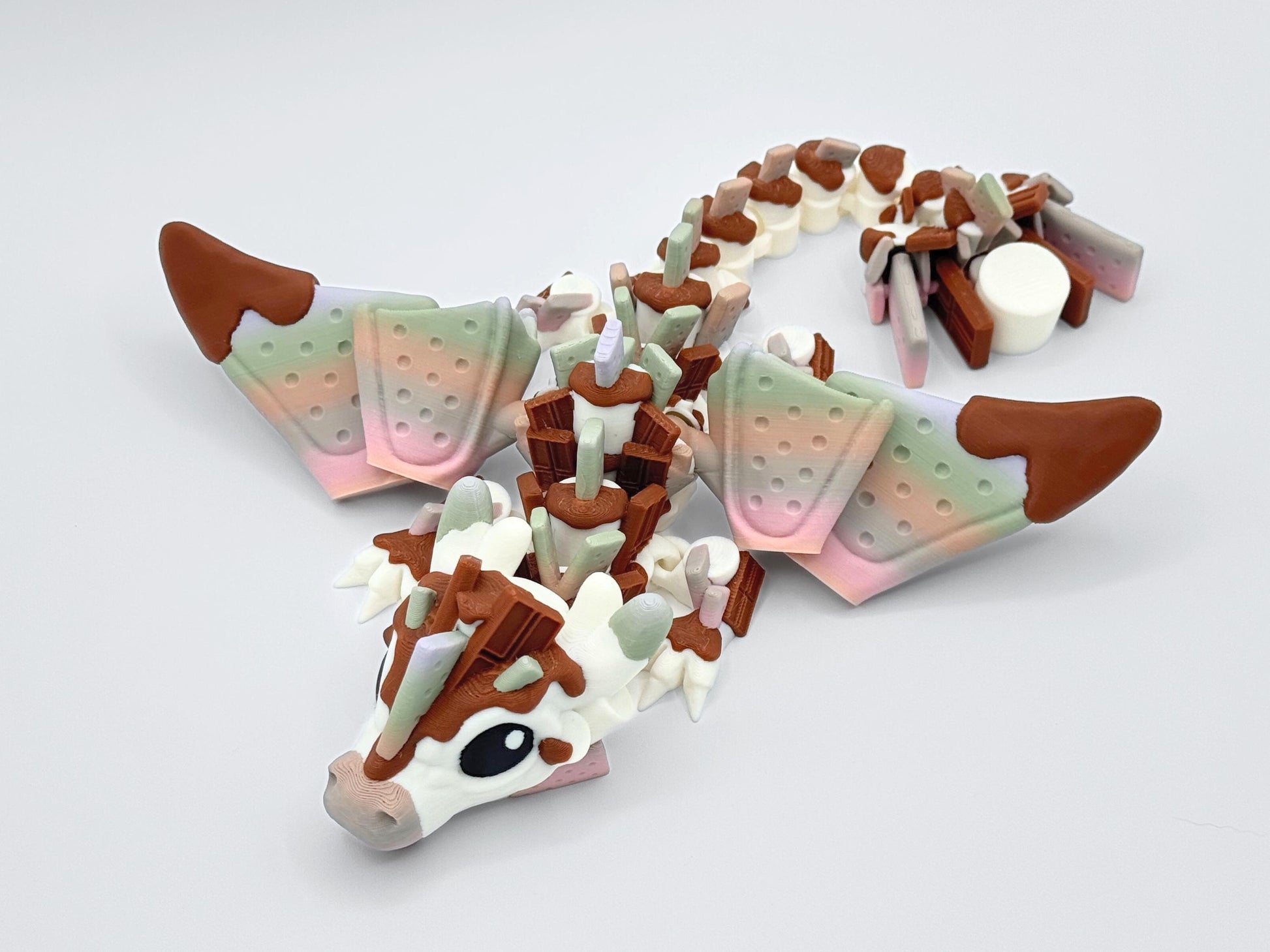 Dynamic top view of S’mores Dragon stretched in an S-shaped pose with graham cracker wings, chocolate horns and marshmallow tail parts visible. The chocolate detailing contrasts with soft gradients on wing panels, white body has marshmallow elements with fidget articulation.
