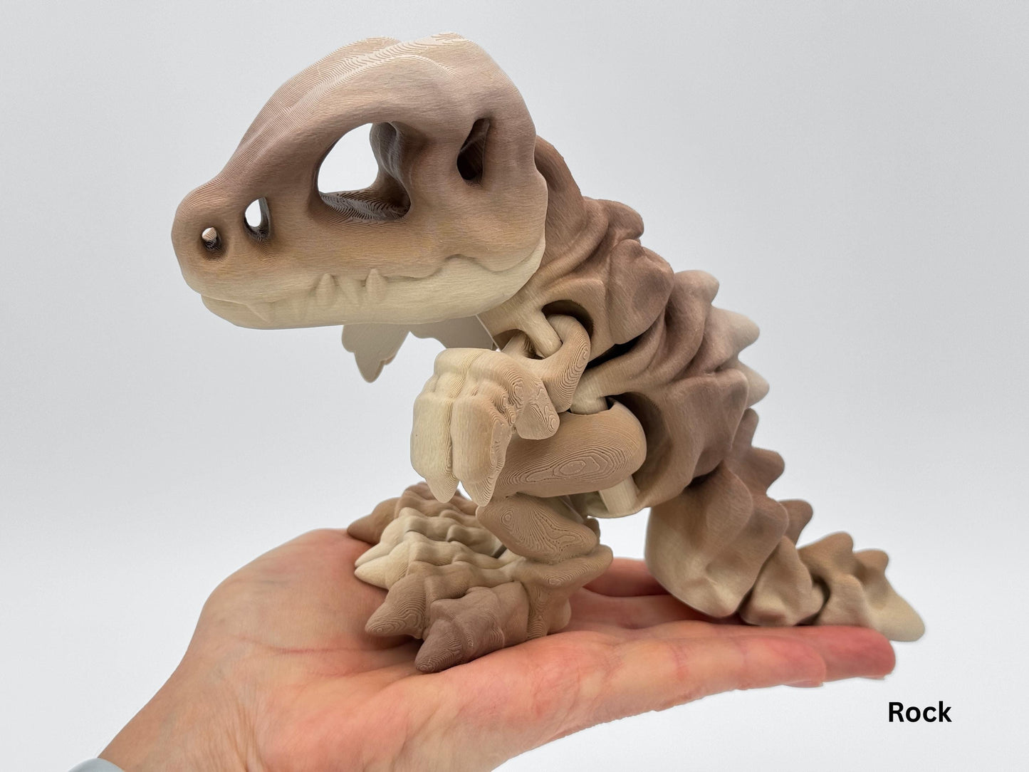 Close-up of the in brown gradient 3D printed articulated dinosaur, shown resting in the palm of a hand for size reference. The figurine has flexible segments, a large hollow eye socket skull, and clawed feet.