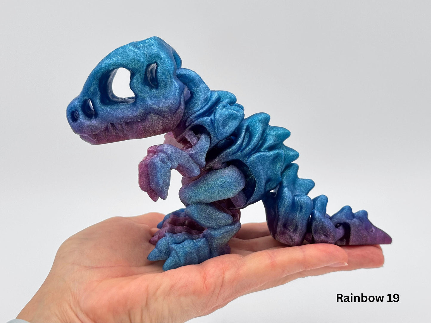 Articulated dinosaur fidget in shimmering rainbow blue, purple and pink gradient filament, displayed in the palm of a hand. The 3D printed T-rex style dragon has flexible joints, short arms, and a segmented tail.