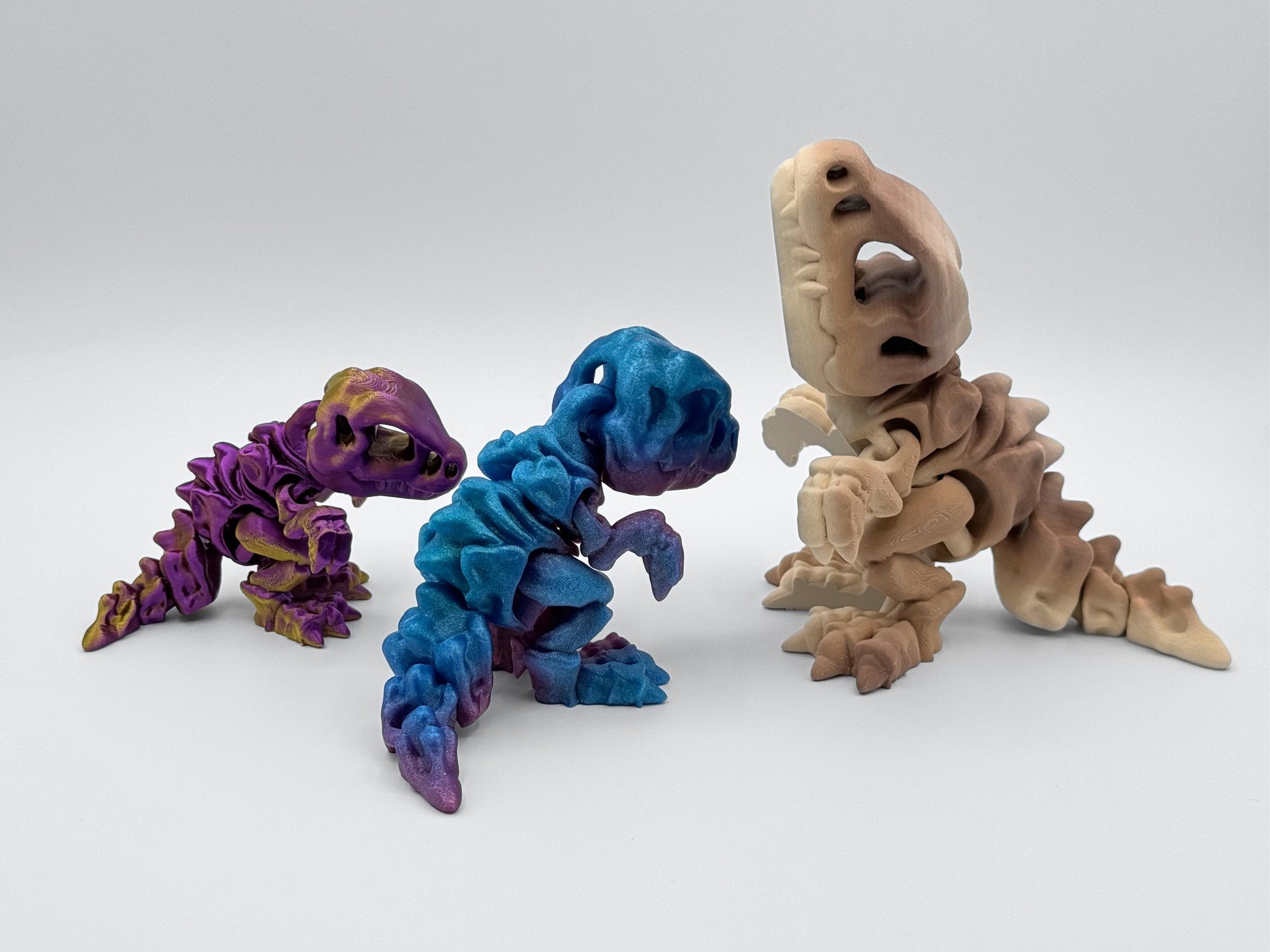 Group of three articulated dinosaur fidgets facing each other, in gold purple, rainbow gradient and tan brown gradient filaments. The 3D printed figurines have jointed bodies, large skull heads, and movable tails.