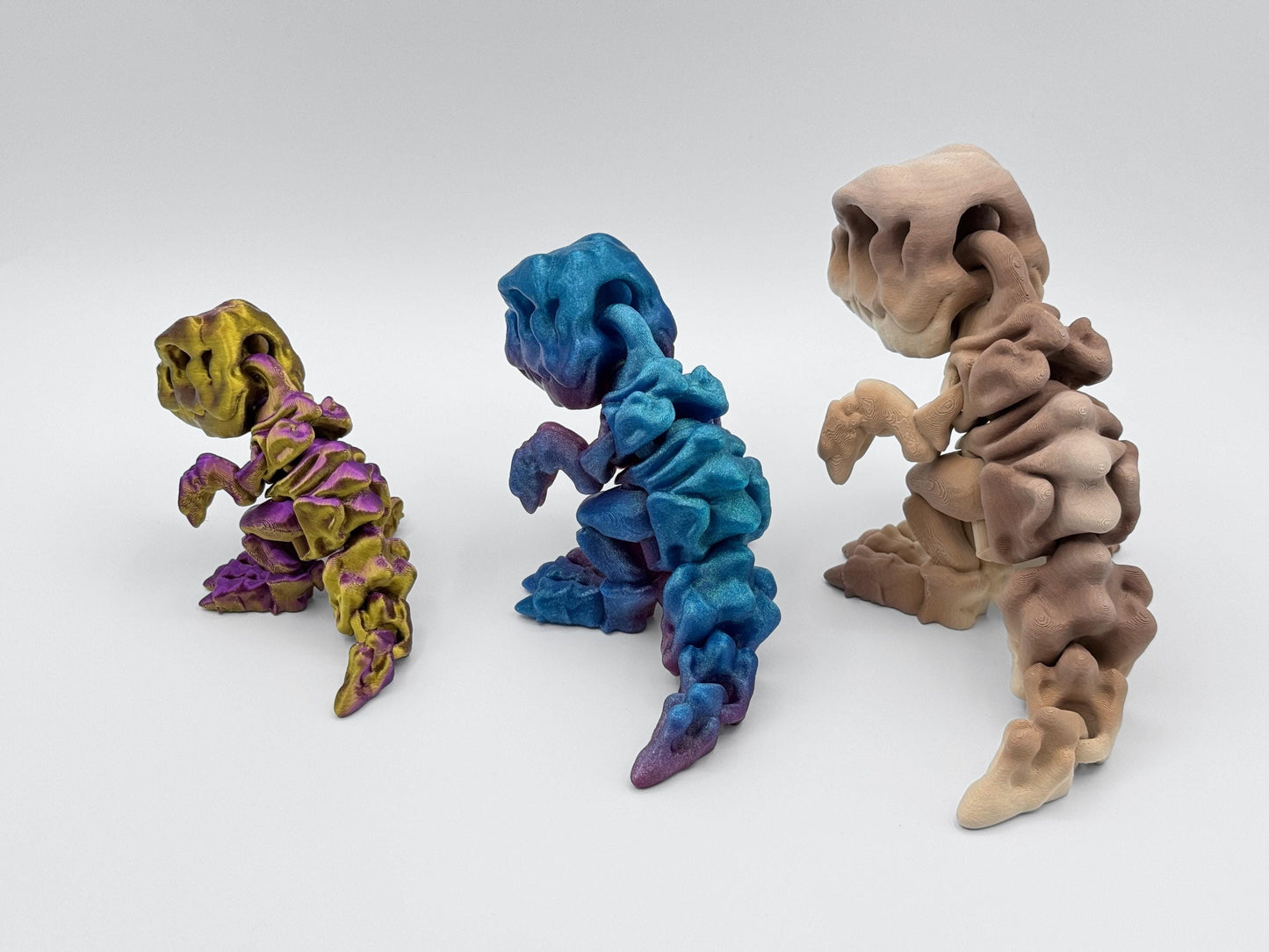 Back view of three 3D printed articulated dinosaurs in gold-purple, rainbow gradient and tan brown. The jointed tails and body segments are visible, showing flexible movement and sculpted texture.
