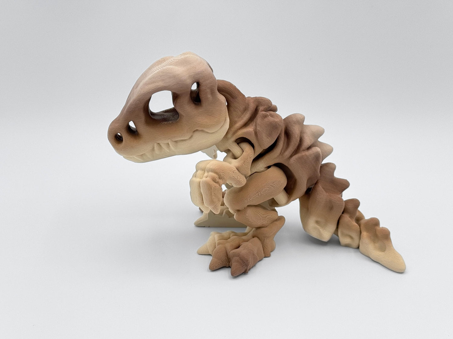 3D printed articulated dinosaur figurine in brown gradient, posed standing on a flat surface. The small T-rex style dragon shows detailed joints, a large skull head, short arms, and a curled tail.