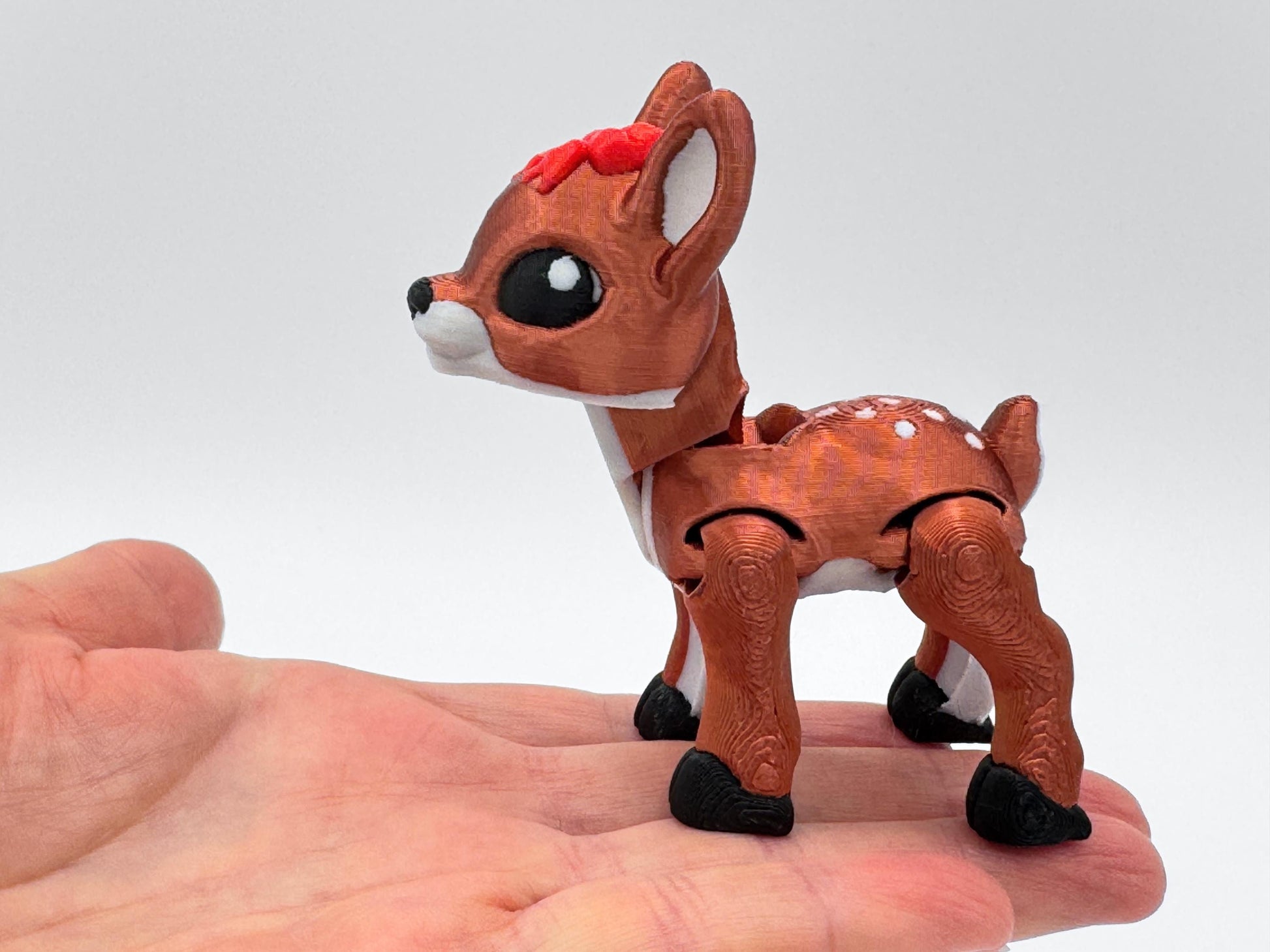 Close-up of a copper red articulated reindeer figurine with a red bow on its head, shown resting in a hand to demonstrate scale. The reindeer has white accents, black hooves, and large eyes, with flexible joints. This Christmas reindeer charm works as a fidget accessory, seasonal collectible, or holiday decoration.
