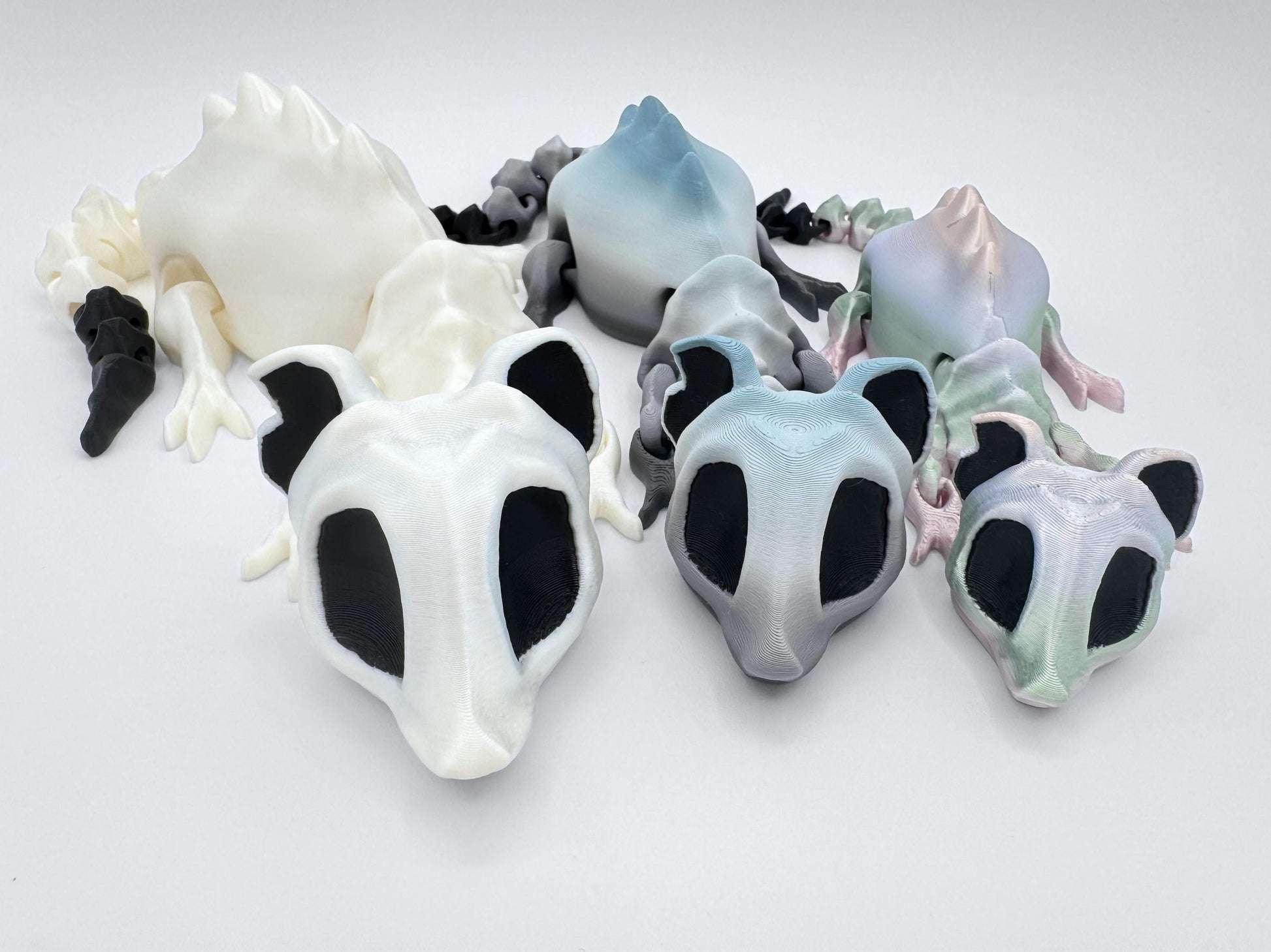 Three 3D printed articulated bone rat figurines in different colors and sizes are arranged side-by-side on a white background. The front row shows their skeletal-style heads with large black eye sockets, while the back row reveals their segmented bodies and tails. Colors include white with black accents, gray gradient, and pastel rainbow gradient.