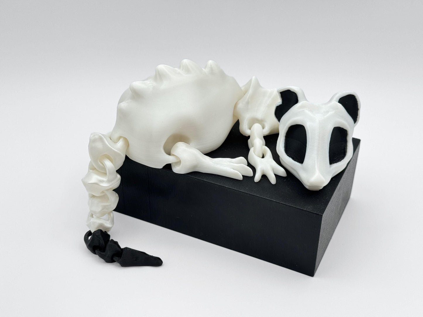 Angled view of 3D printed articulated bone rat figurine in white with black accents, lying on a black block. The tail drapes off the side while the head faces forward, emphasizing the smooth segmented joints.