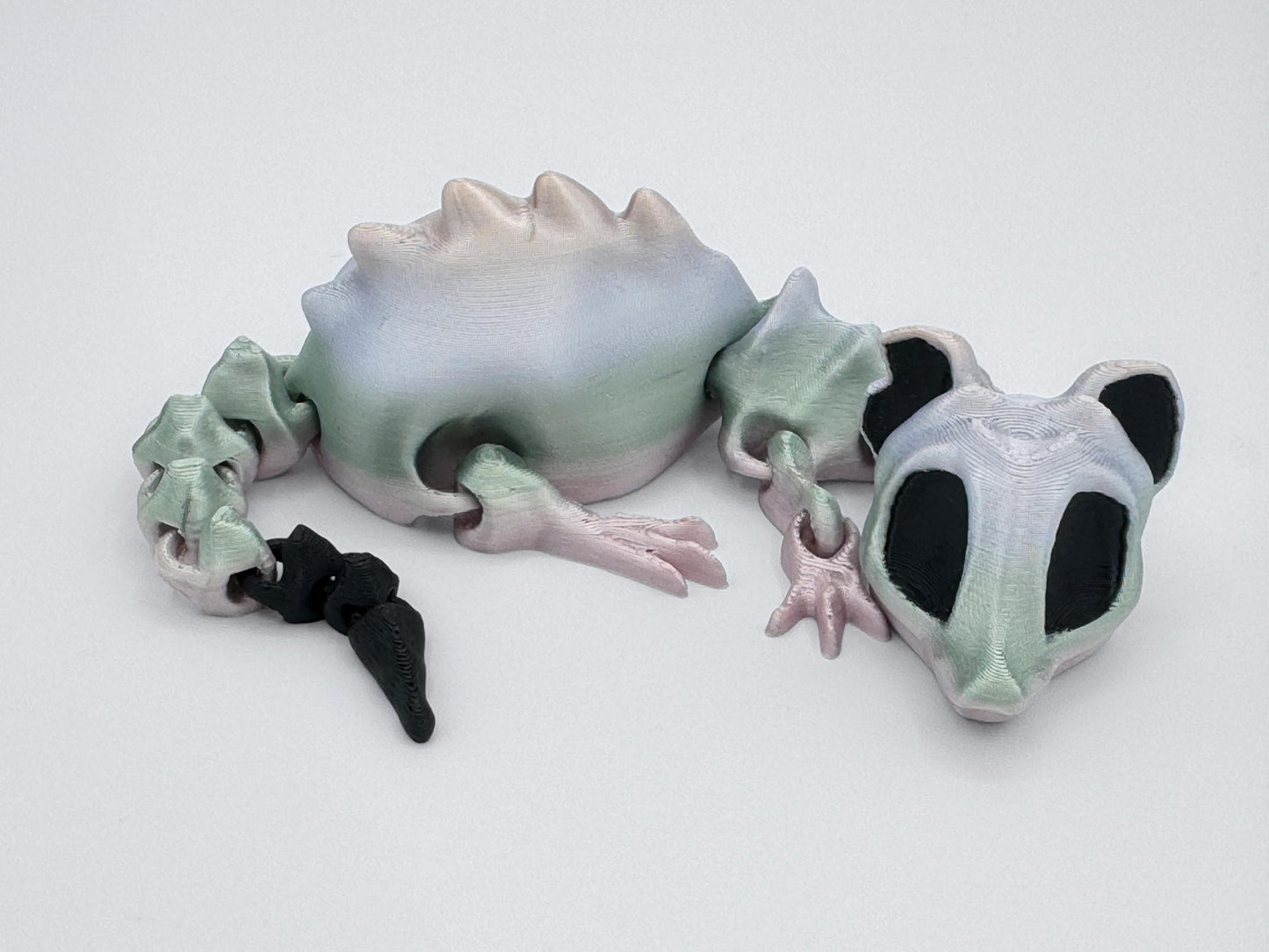 A pastel 3D printed articulated bone rat figurine lying on a white surface. Its segmented body is shaded in soft gradients of green, pink, and pale white, with a black tail tip. Large black eye sockets give it a skeletal, slightly spooky look. The smooth, jointed design allows the head, legs, and tail to move for posing or display.