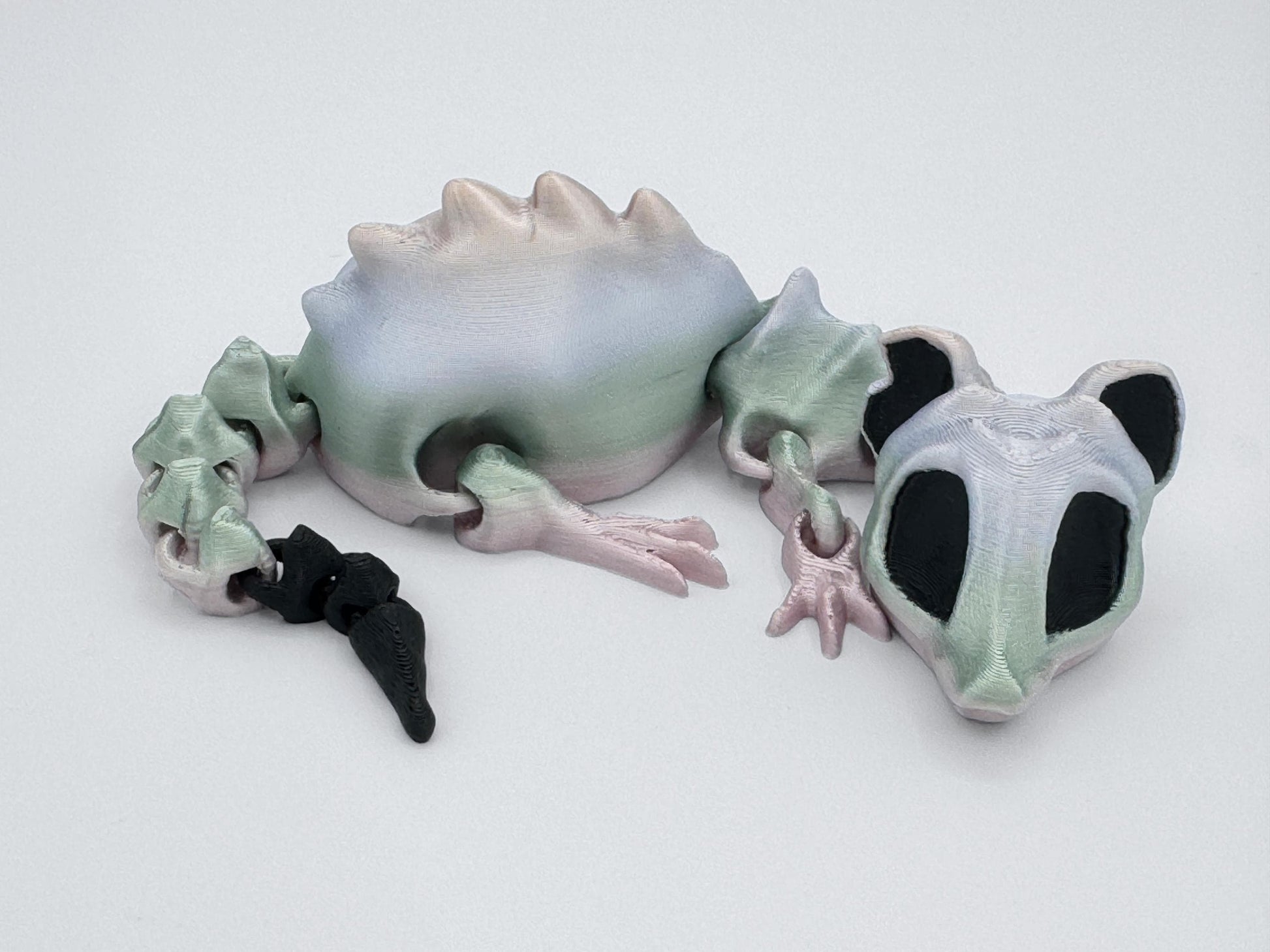 A pastel 3D printed articulated bone rat figurine lying on a white surface. Its segmented body is shaded in soft gradients of green, pink, and pale white, with a black tail tip. Large black eye sockets give it a skeletal, slightly spooky look. The smooth, jointed design allows the head, legs, and tail to move for posing or display.