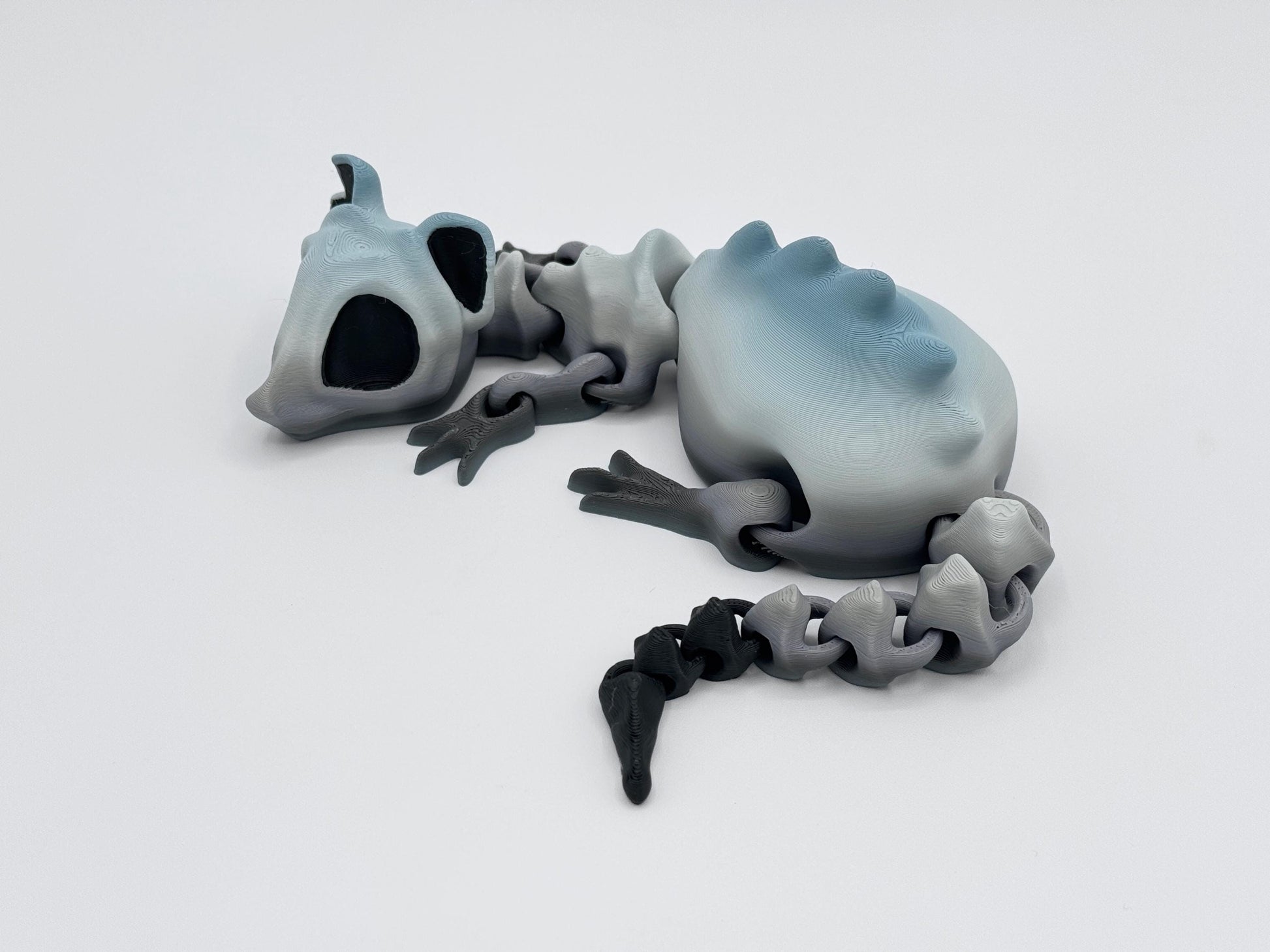 3D Printed articulated bone rat figurine posed on a white surface. Its body fades from light gray to a soft blue-gray, with a black tail tip and smooth, connected segments forming the spine. The head features large black eye sockets for a skeletal effect. The jointed tail and limbs are positioned naturally to show articulation.