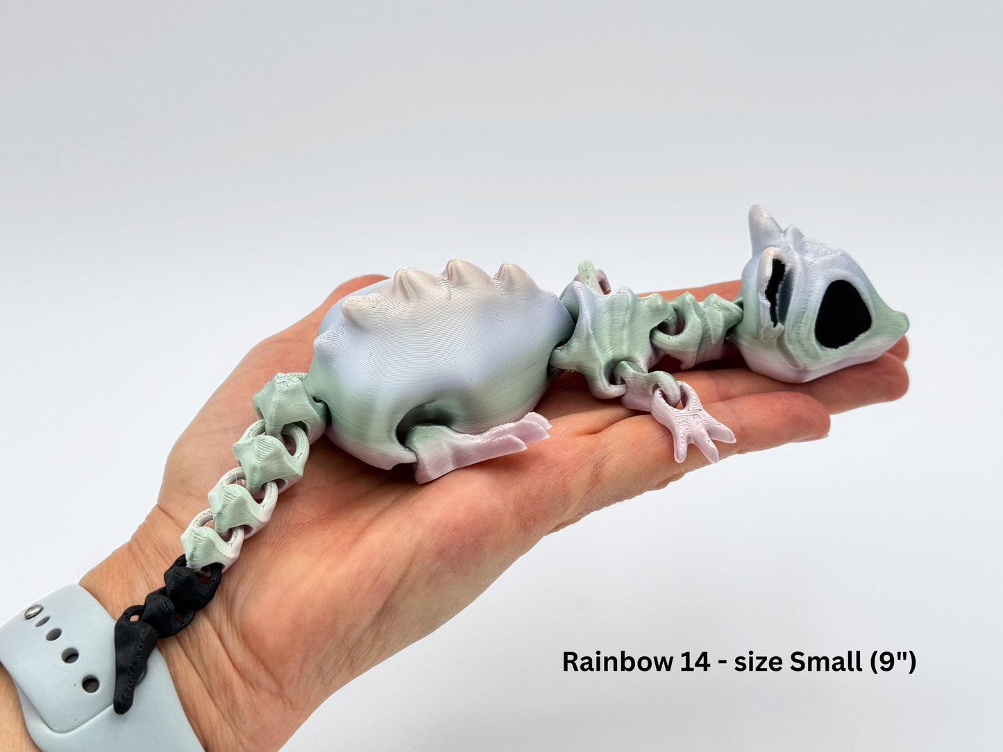 A small 9-inch articulated bone rat figurine rests across an adult hand, showing its full length from nose to tail. The body is printed in soft pastel shades of green, pink, and white with a smooth, segmented design that allows the legs, tail, and head to move. The tail tip is black, adding contrast to the light, skeletal-inspired body. The photo provides a clear size reference against the hand and watch band.