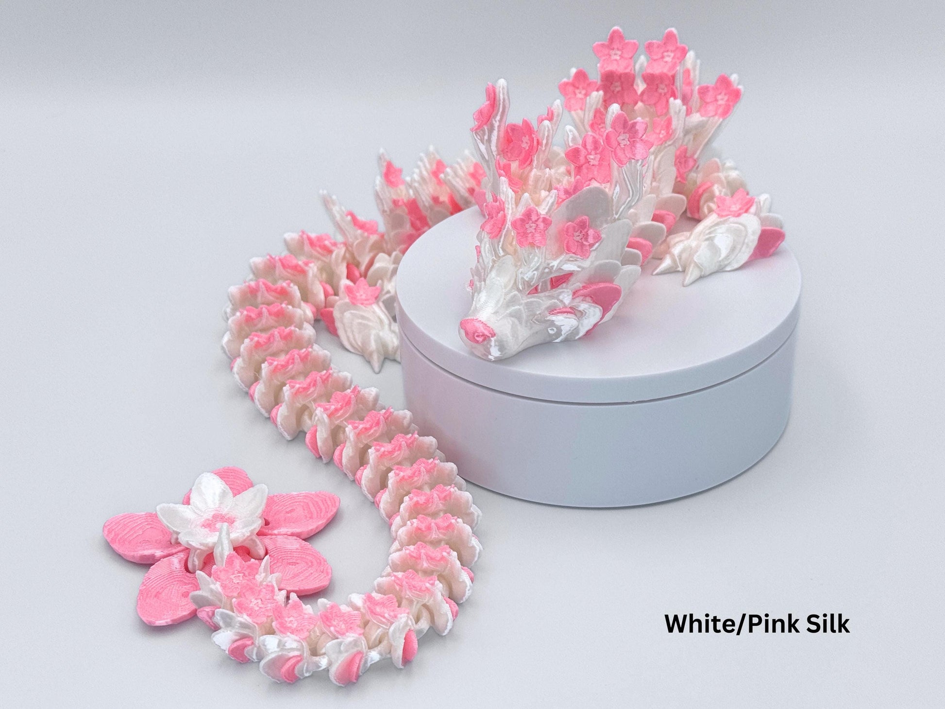 Articulated Cherry Blossom dragon figurine in white and pink silk, displayed on a round stand. The 3D printed dragon shows blossom textures across the head, wings, and tail with flowing segmented joints.