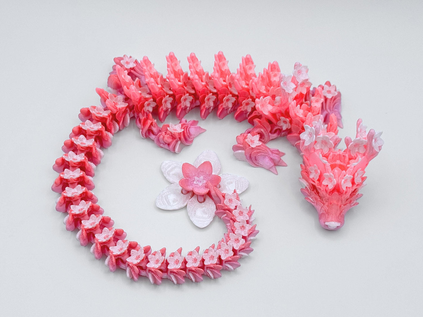 Top view of a pink and peach 3D printed dragon with flower-shaped scales and a long segmented body curled into an S shape, ending in a large blossom tail.