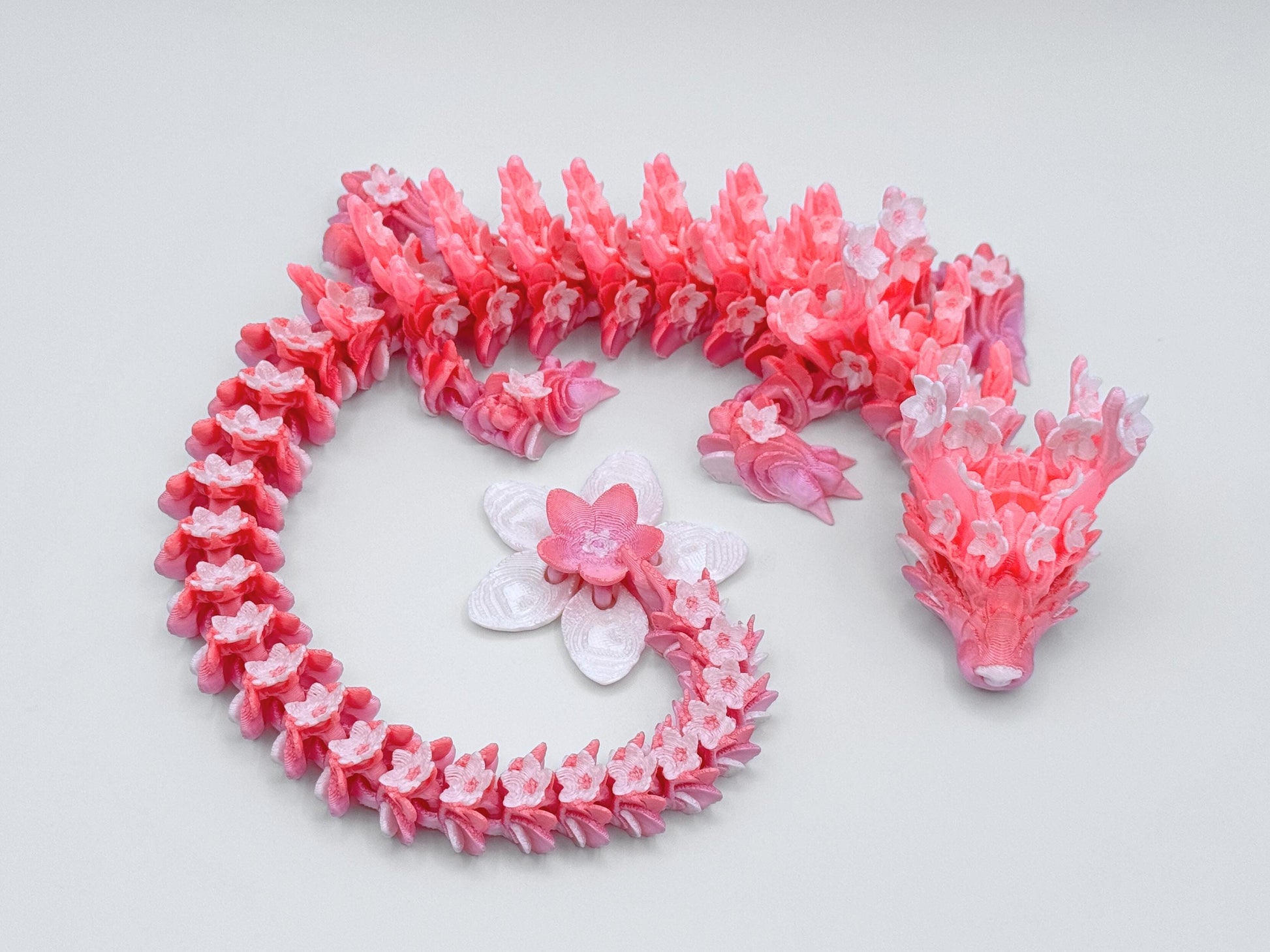 Top view of a pink and peach 3D printed dragon with flower-shaped scales and a long segmented body curled into an S shape, ending in a large blossom tail.